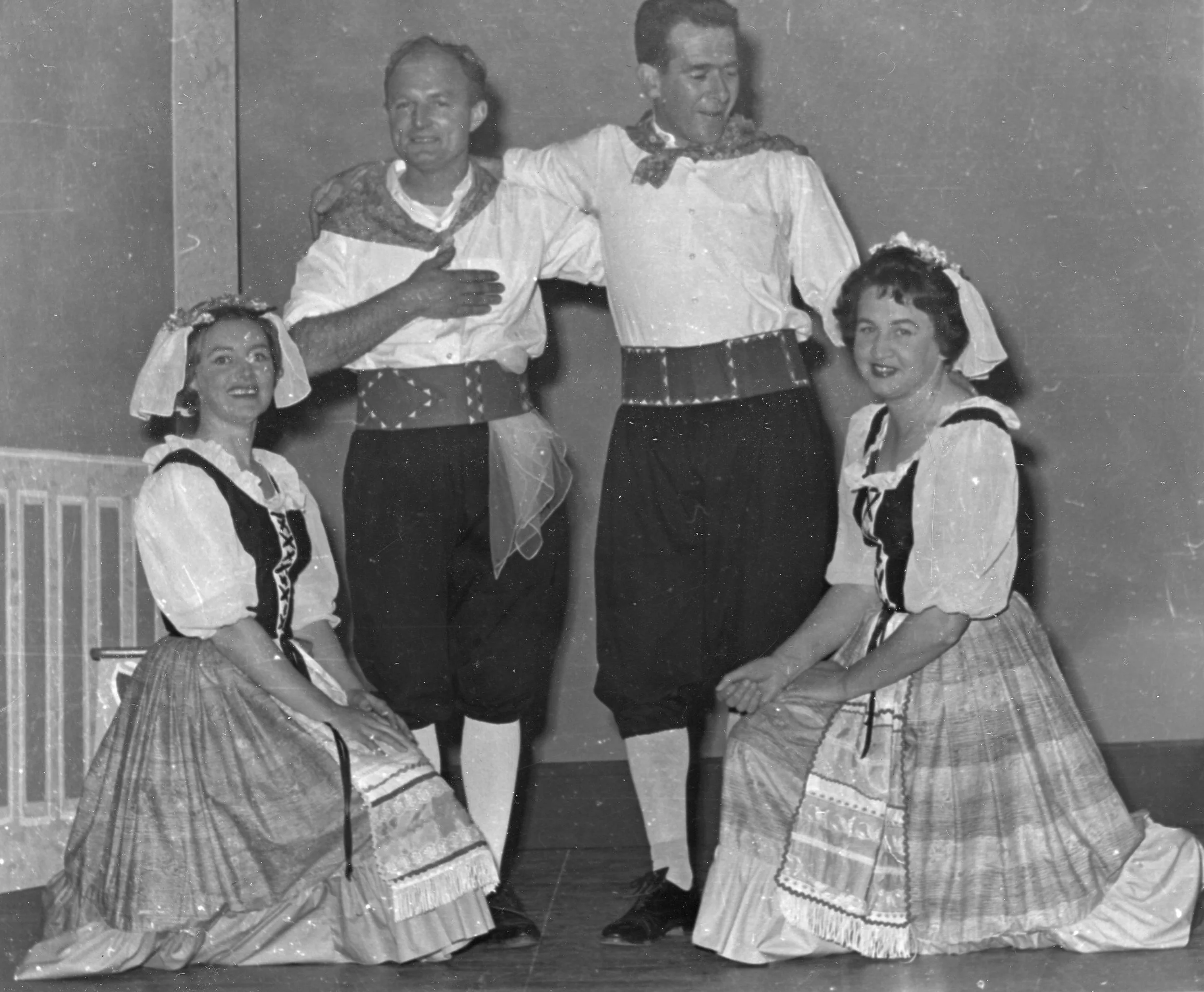 Black and white photo of four performers in traditional European folk costumes. Two women are kneeling in front, wearing long skirts, blouses, and headscarves. Two men are standing behind them, in shirts with rolled-up sleeves, wide belts, and dark p