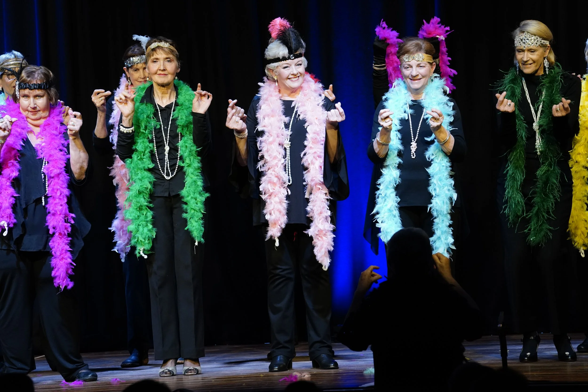 Cowra Theatre Company Group of senior women on stage dressed in black, wearing colorful feather boas and headbands, performing in a show.