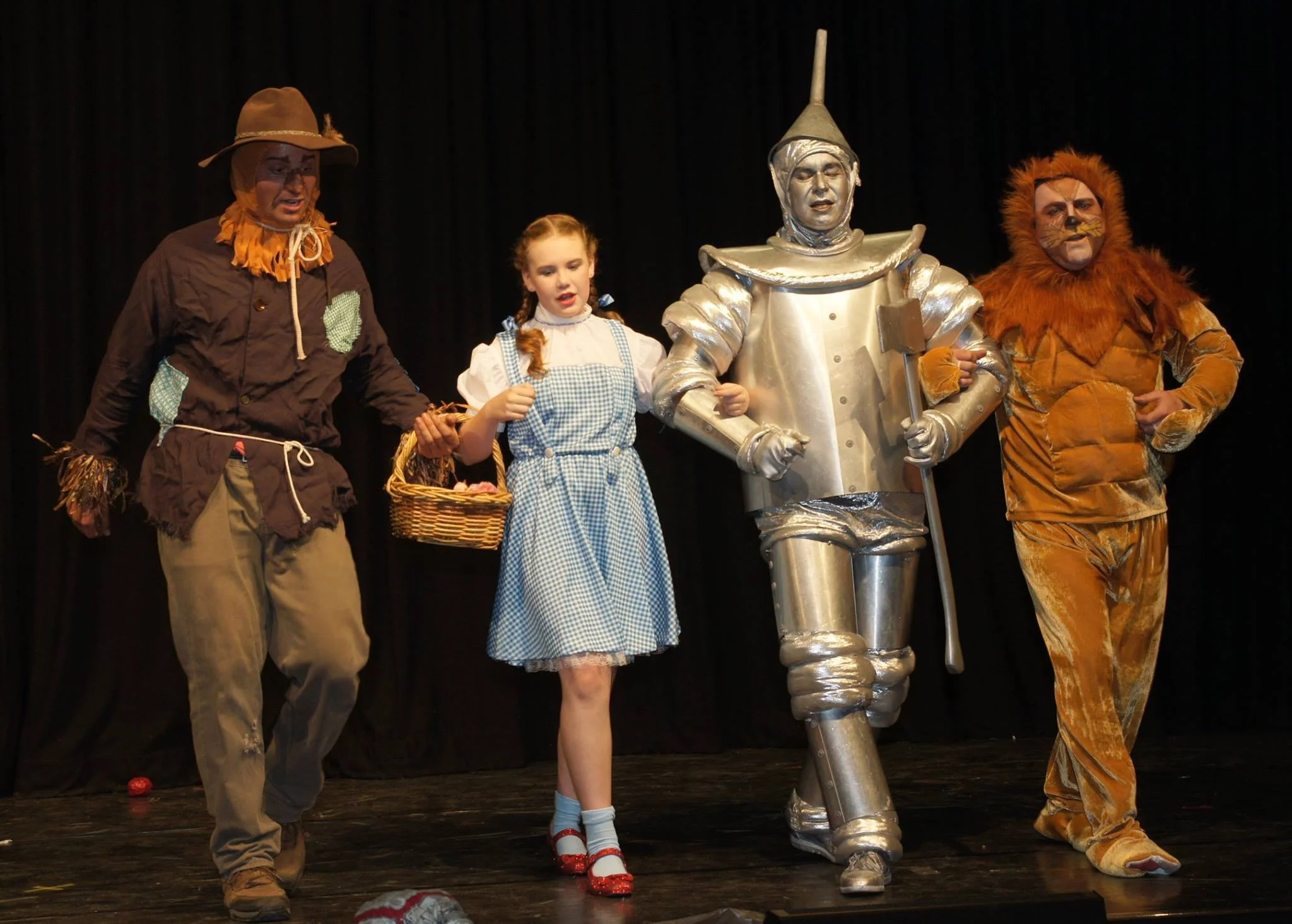 Children and actors dressed as characters from 'The Wizard of Oz' on stage, including the Scarecrow, Dorothy, Tin Man, and Cowardly Lion.