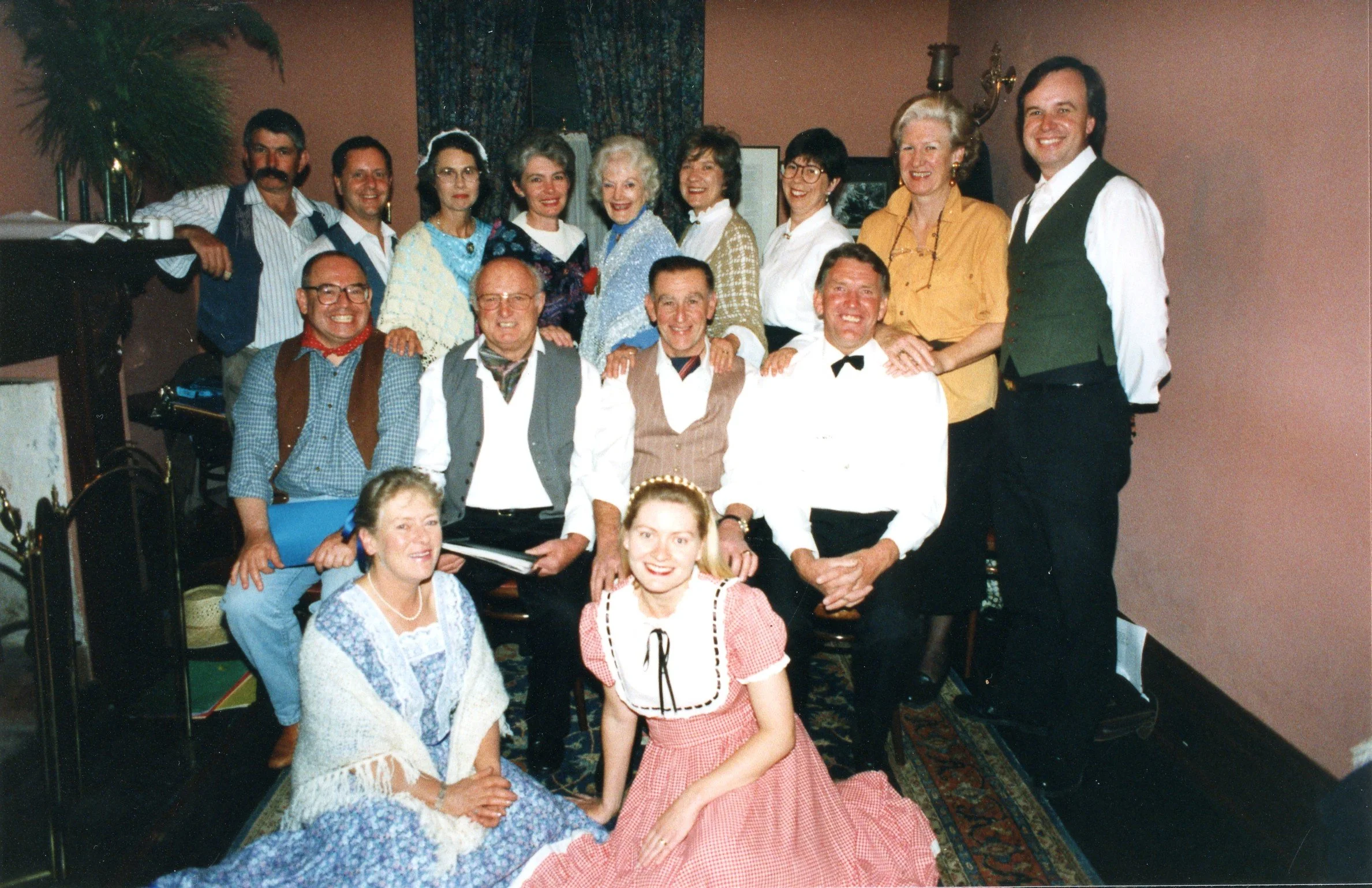 Group of people dressed in vintage or period costumes, gathered in a room for a theatrical or historical reenactment, with some standing and others seated, smiling at the camera.