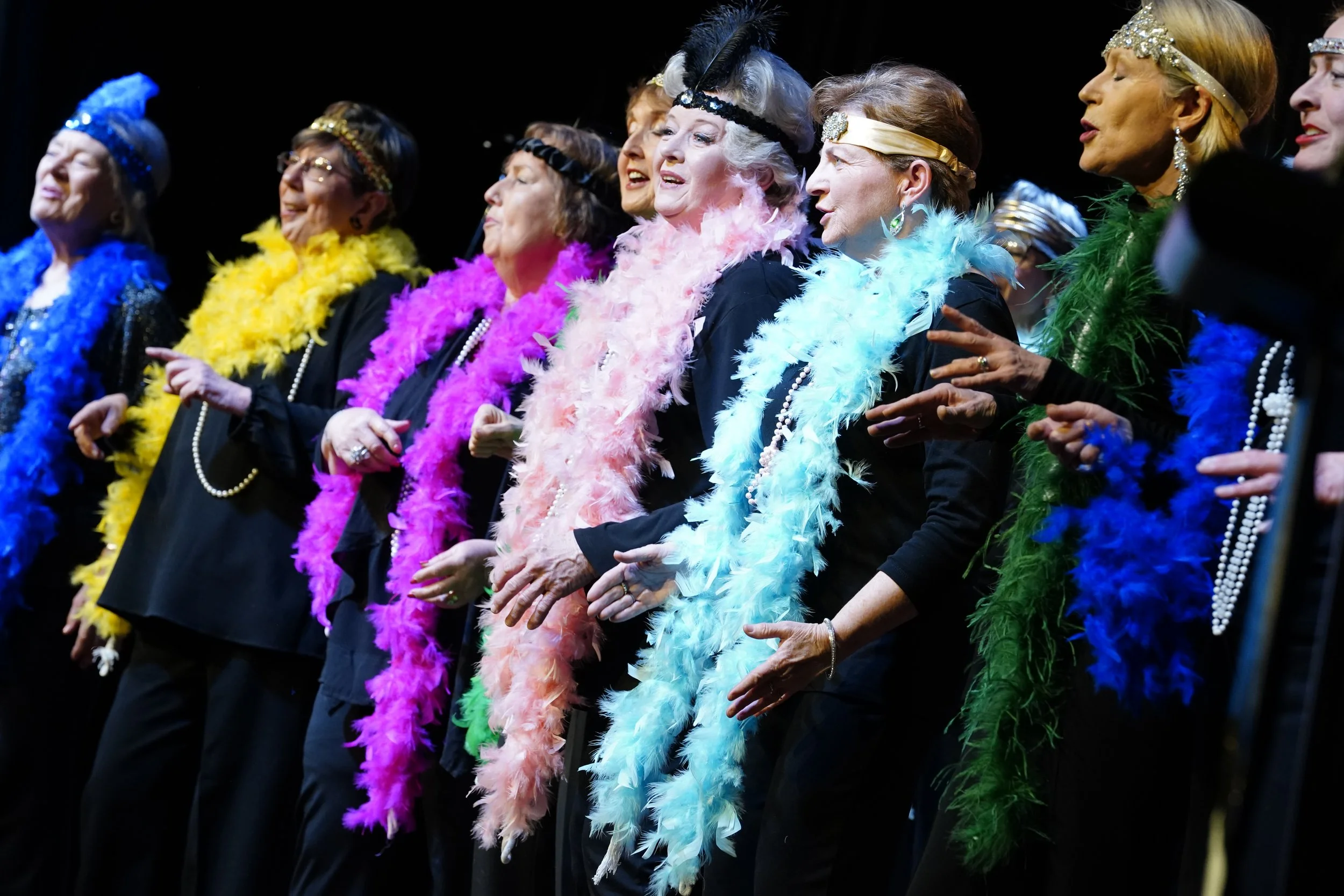 A group of older women dressed in black, wearing colorful feather boas and pearl necklaces, performing on stage.