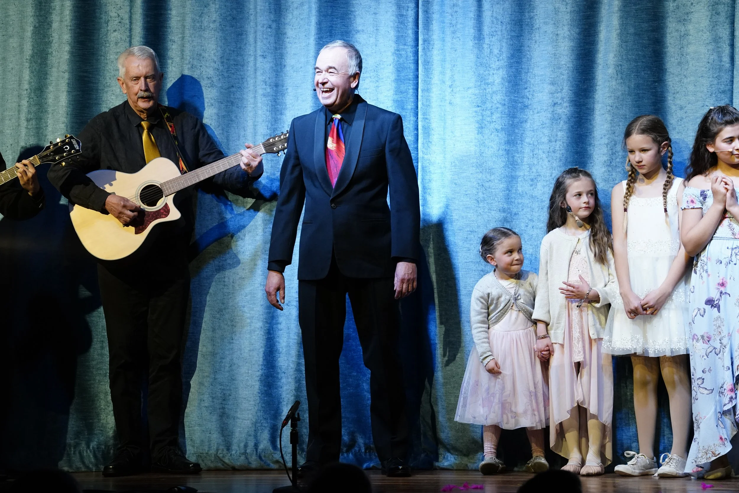 A man playing guitar on stage with a group of children, some holding hands, standing in front of blue curtains, with another man laughing nearby.