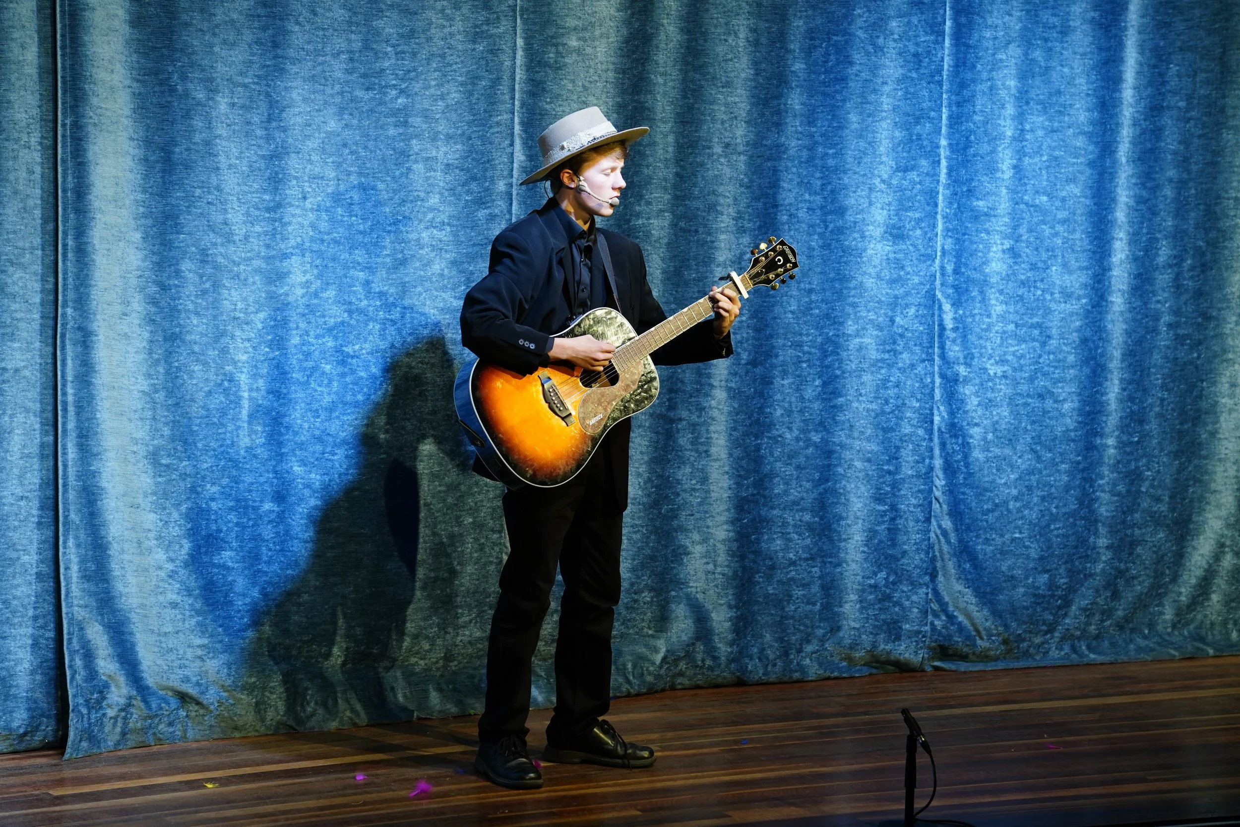 Person playing guitar on stage, wearing a gray hat and black clothing, with a blue curtain backdrop.