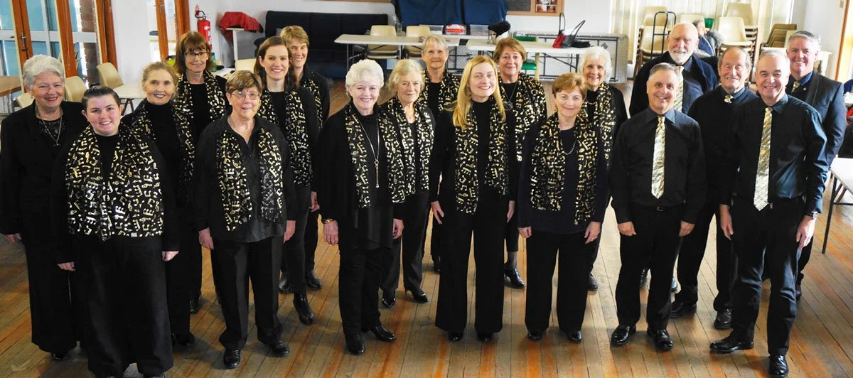 Group of fifteen people, mostly women, dressed in black with matching black and gold patterned scarves and ties, standing in a room with wood flooring, chairs, and tables in the background.