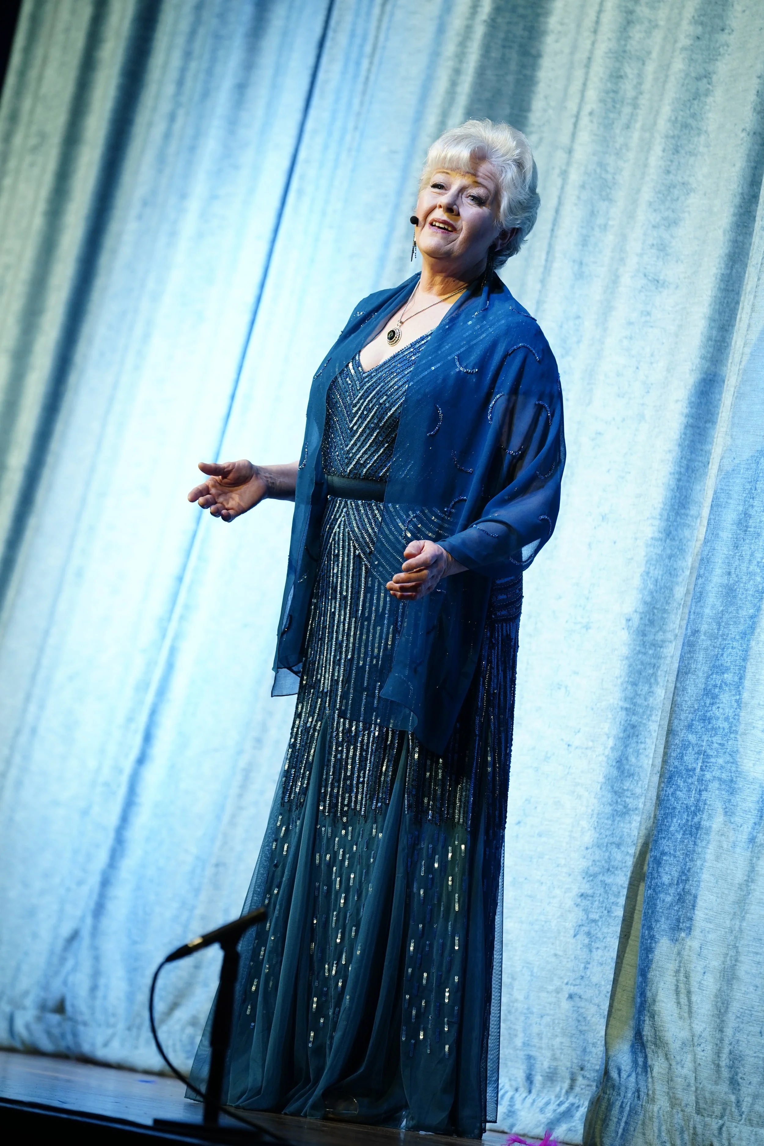 Cowra Theatre Company Older woman performing on stage, wearing a blue, beaded, floor-length gown and matching sheer jacket, with light-colored hair styled in an updo, standing in front of a light blue curtain.