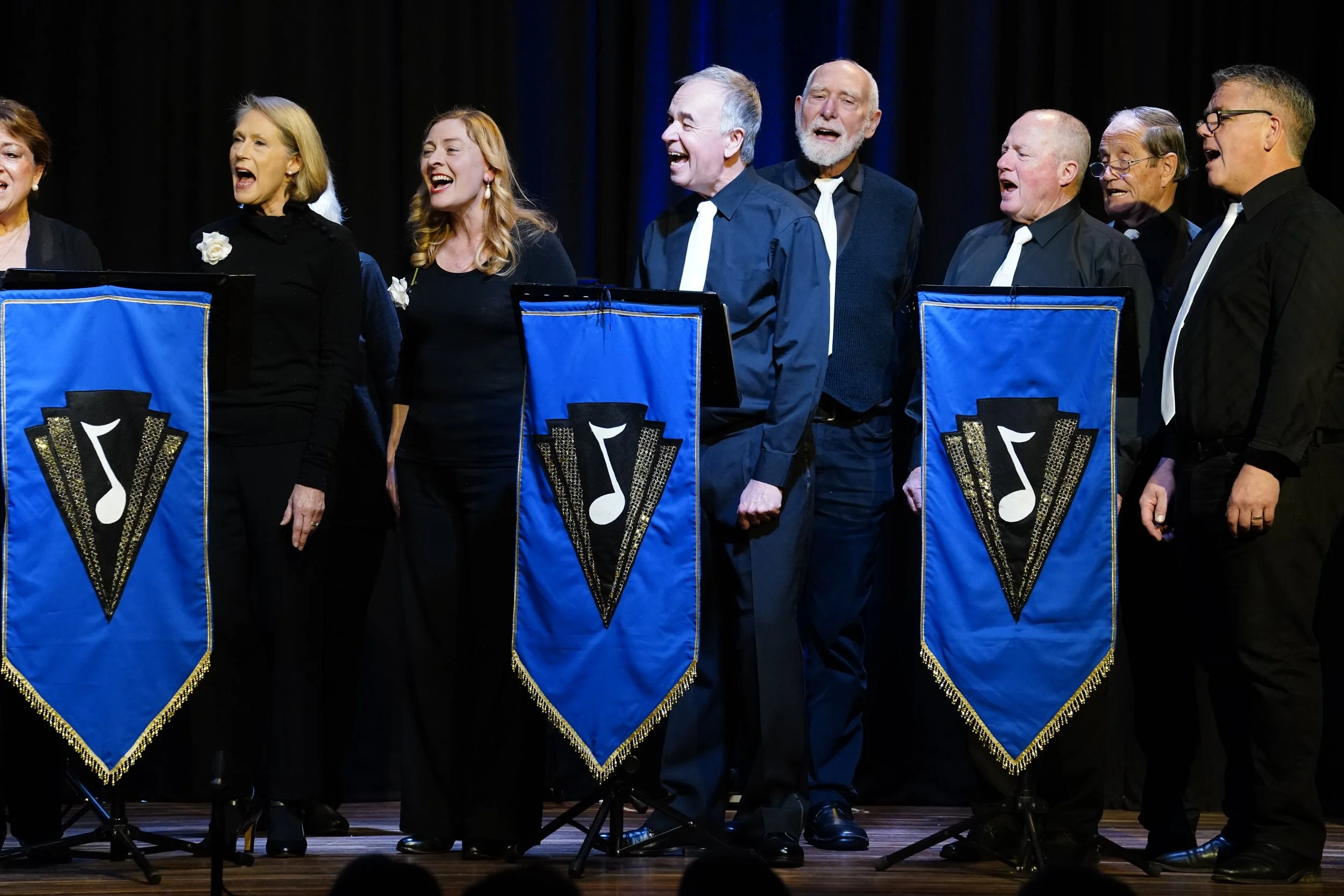Cowra Theatre Company A choir of men and women singing on stage, standing behind blue music stands decorated with a black and white musical note logo.