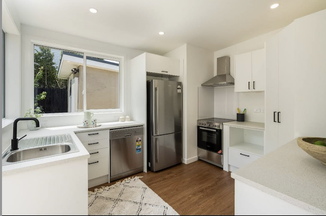 Modern kitchen with white cabinets, stainless steel appliances including a refrigerator, stove, microwave, and dishwasher, a window above the sink, and a wooden floor.