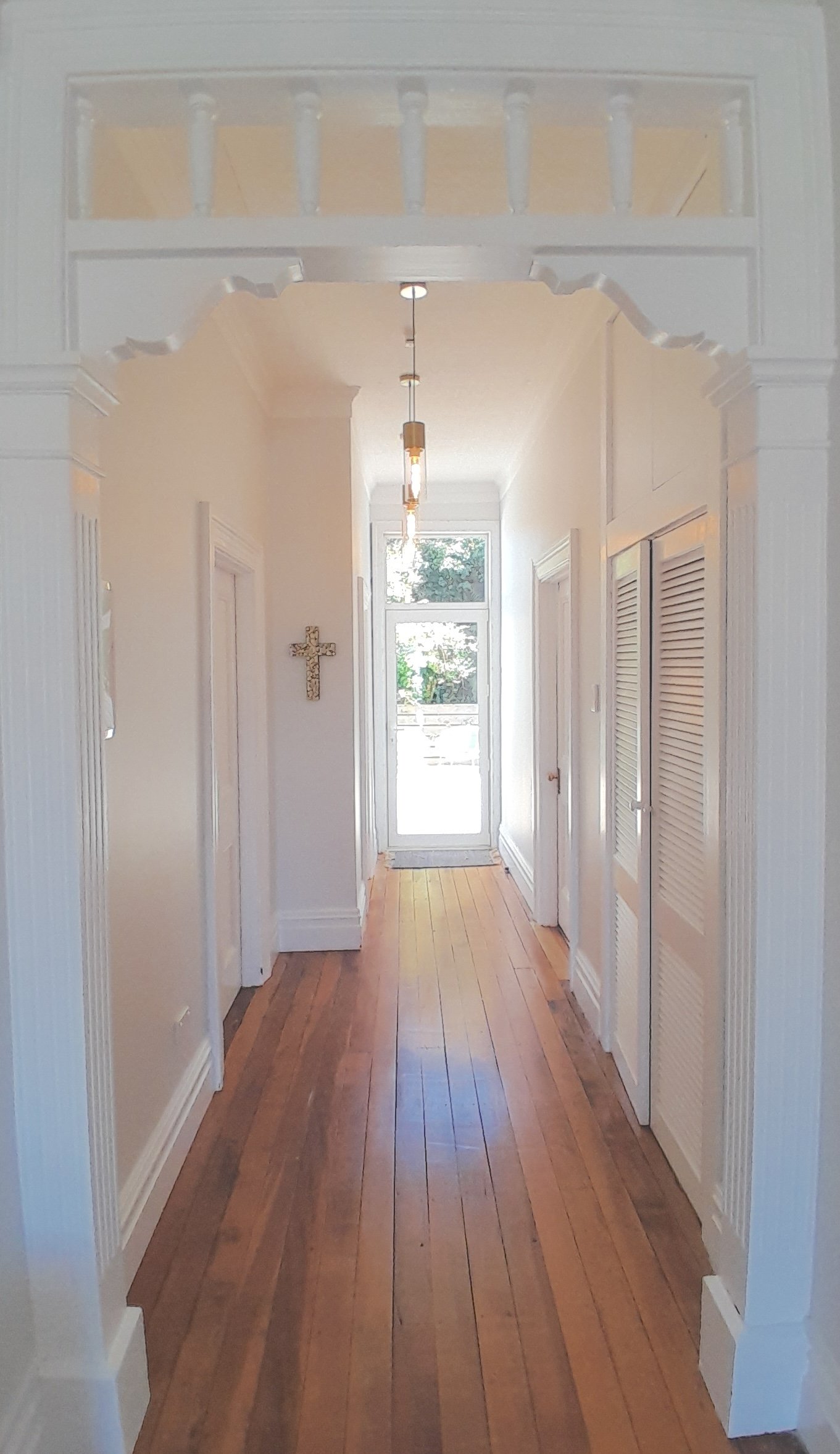 Bright hallway with wooden floor, white walls, and a glass door at the end, illuminated by hanging pendant lights and natural light from outside, with a decorative cross on the wall.