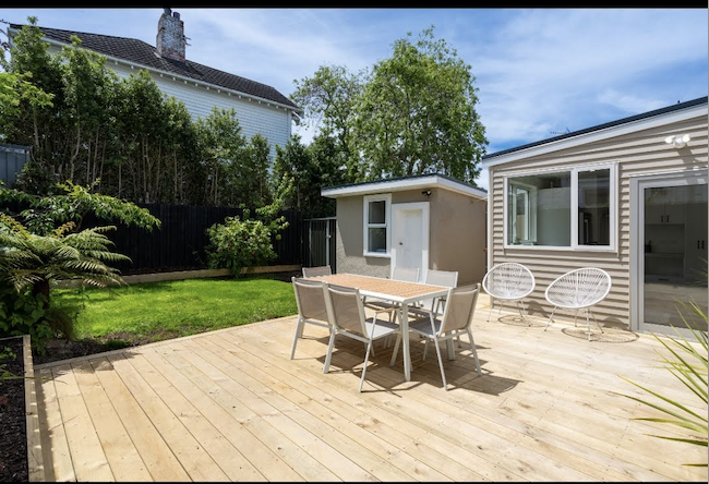 Backyard patio with wooden deck, outdoor dining table, six chairs, garden area, small shed, two white circular chairs, and a house with large windows.