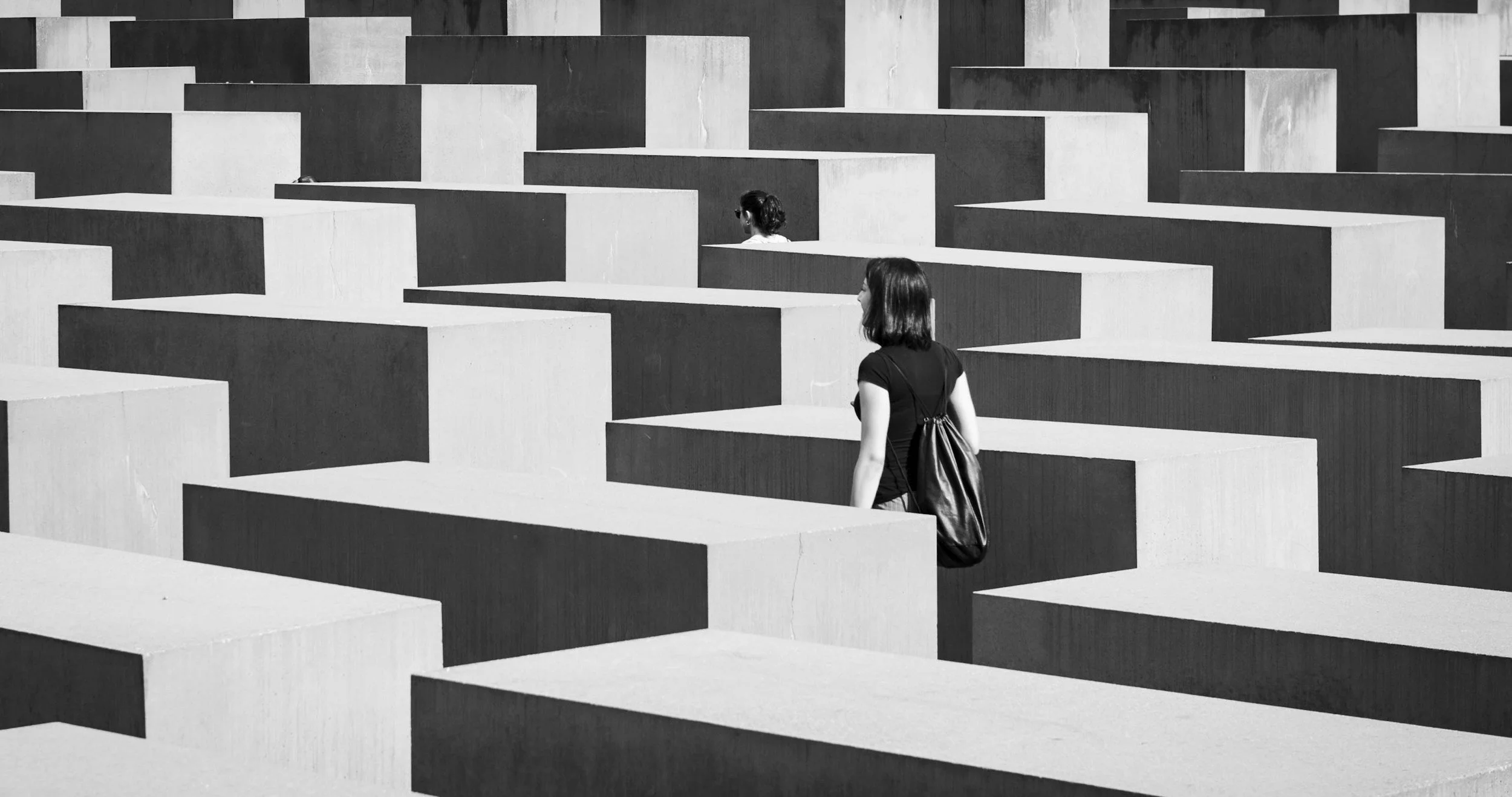 Two women walking among large, maze-like concrete blocks in an outdoor setting, captured in black and white.