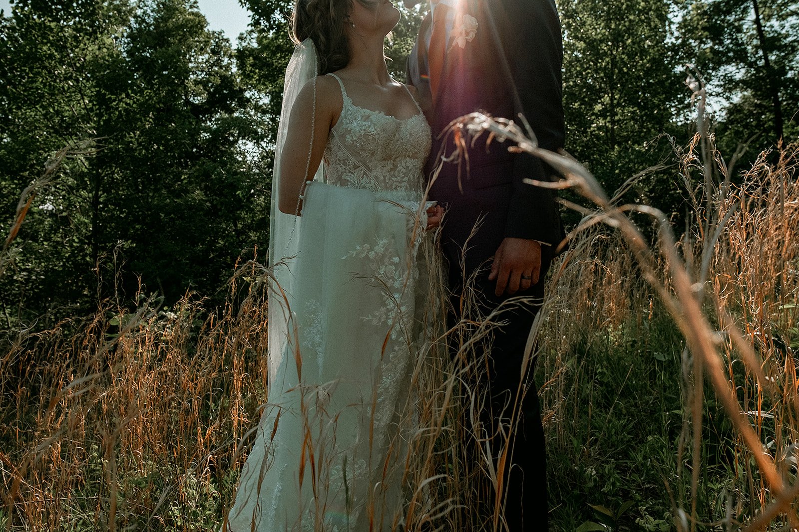 A bride and groom holding hands in a grassy outdoor setting with sunlight filtering through trees.