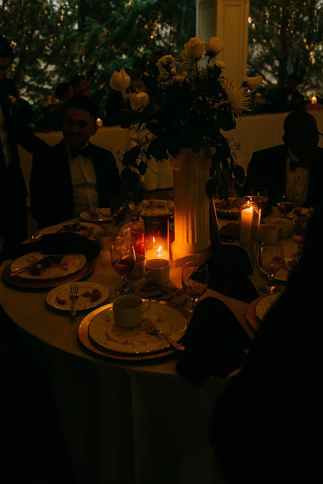 A dimly lit dinner table with a tall floral centerpiece, lit candles, and guests dressed in formal attire.
