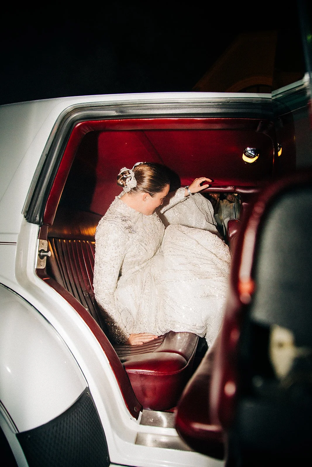 A bride in a white lace wedding dress sitting inside a vintage car with a red leather interior.