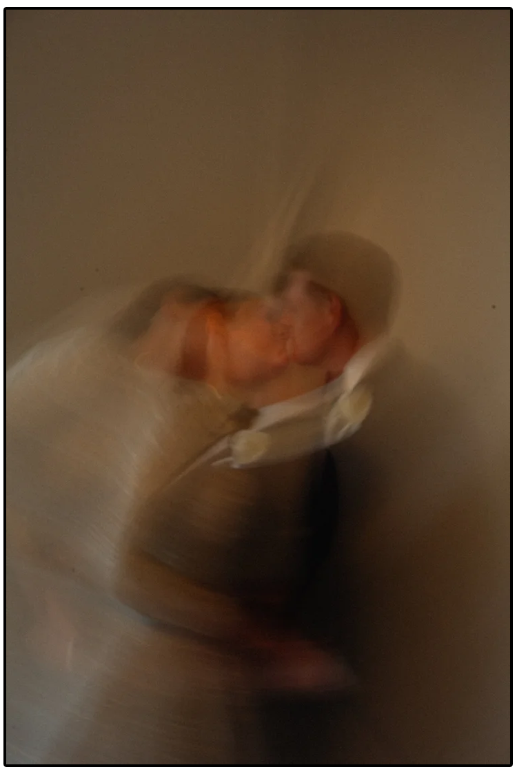 Blurred image of two people kissing, one wearing a hat and the other with short hair, against a plain background.