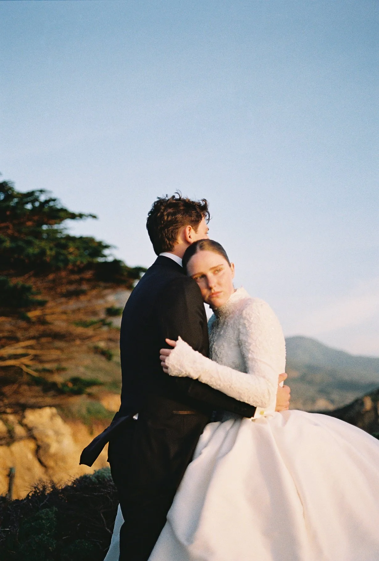 A bride and groom embrace outdoors during sunset. The bride, in a white lace wedding dress, rests her head on the groom’s shoulder. The groom, in a black suit, holds the bride. Background features mountains and a clear blue sky.