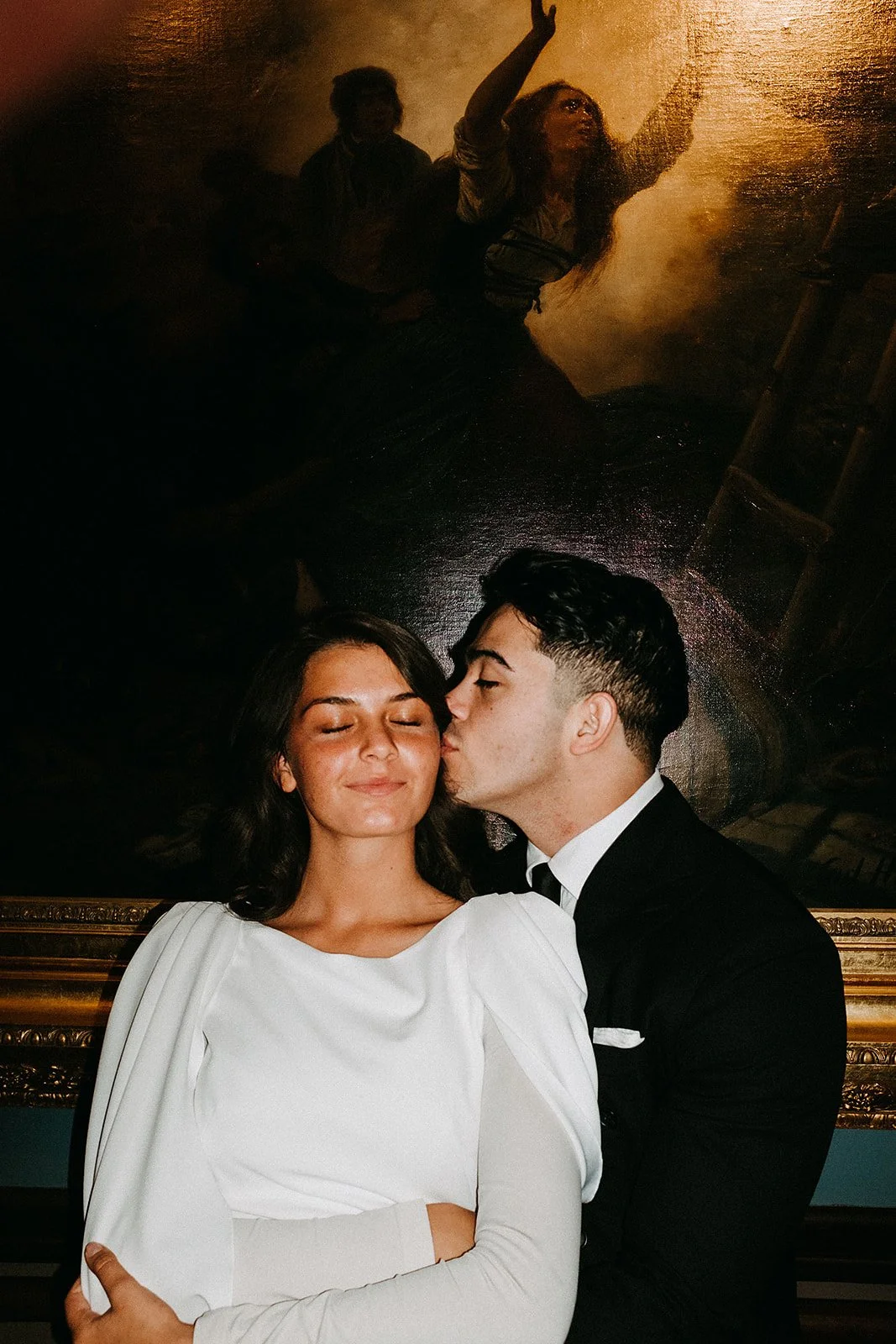 A young couple standing close to each other. The woman has dark hair and is wearing a white dress, with her eyes closed and a smile. The man, dressed in a black tuxedo, is kissing her on the cheek. Behind them is a dark, framed painting of a woman wi