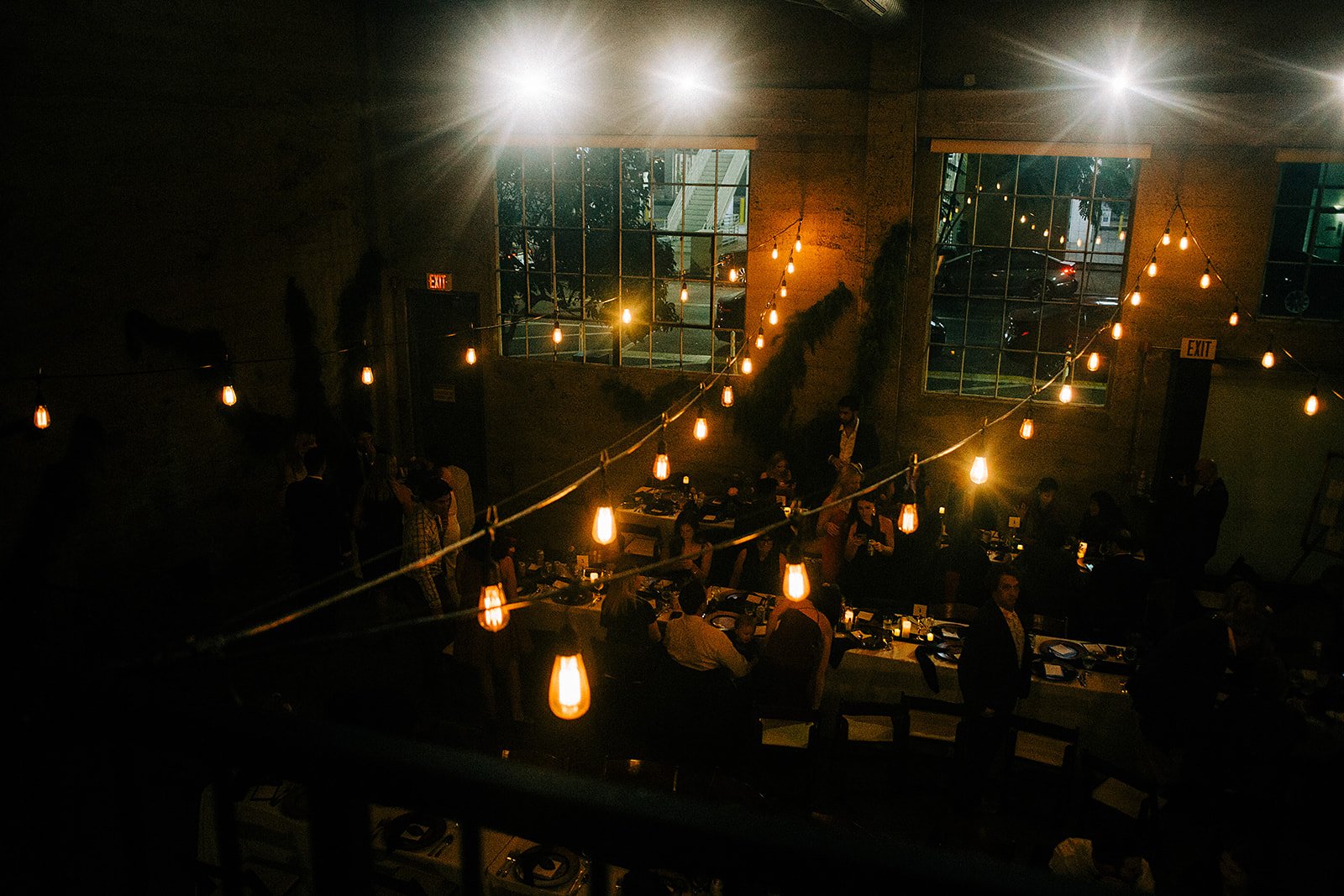 Dimly lit restaurant or event space with hanging string lights, people seated at tables, and large windows showing parked cars outside in the evening.