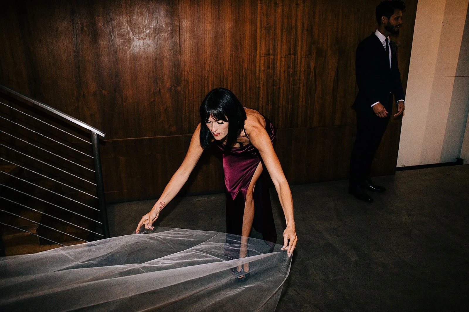 A woman in a dark purple dress bending down to touch a large piece of transparent fabric with a man standing nearby in a dark suit in a room with wood-paneled walls.