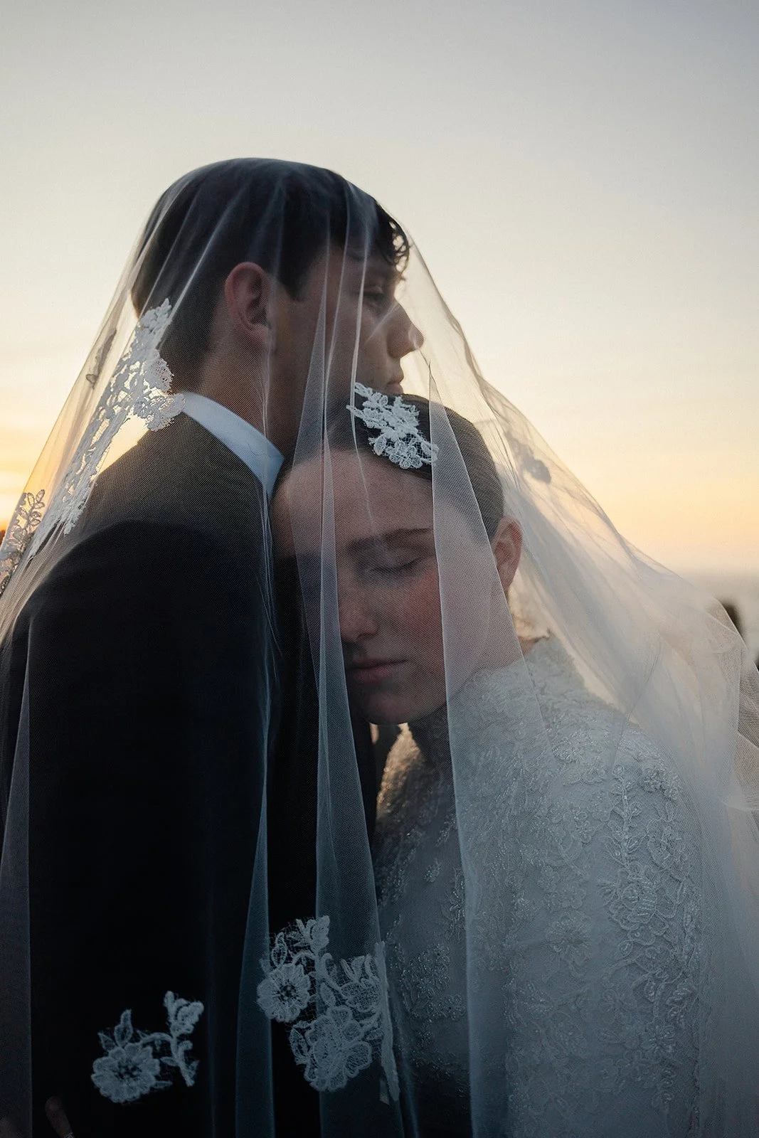 A bride and groom share an intimate moment under a wedding veil during sunset.