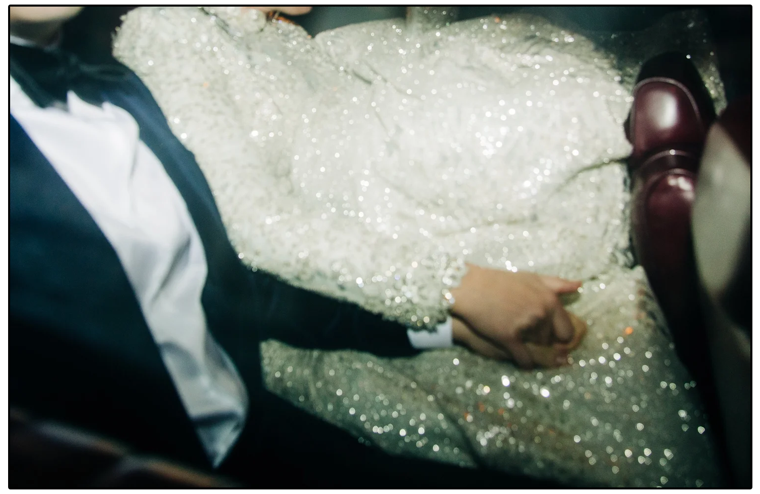 A couple holding hands while sitting together, dressed in wedding attire with the bride wearing a sparkly white dress and the groom in a tuxedo, inside a vehicle.