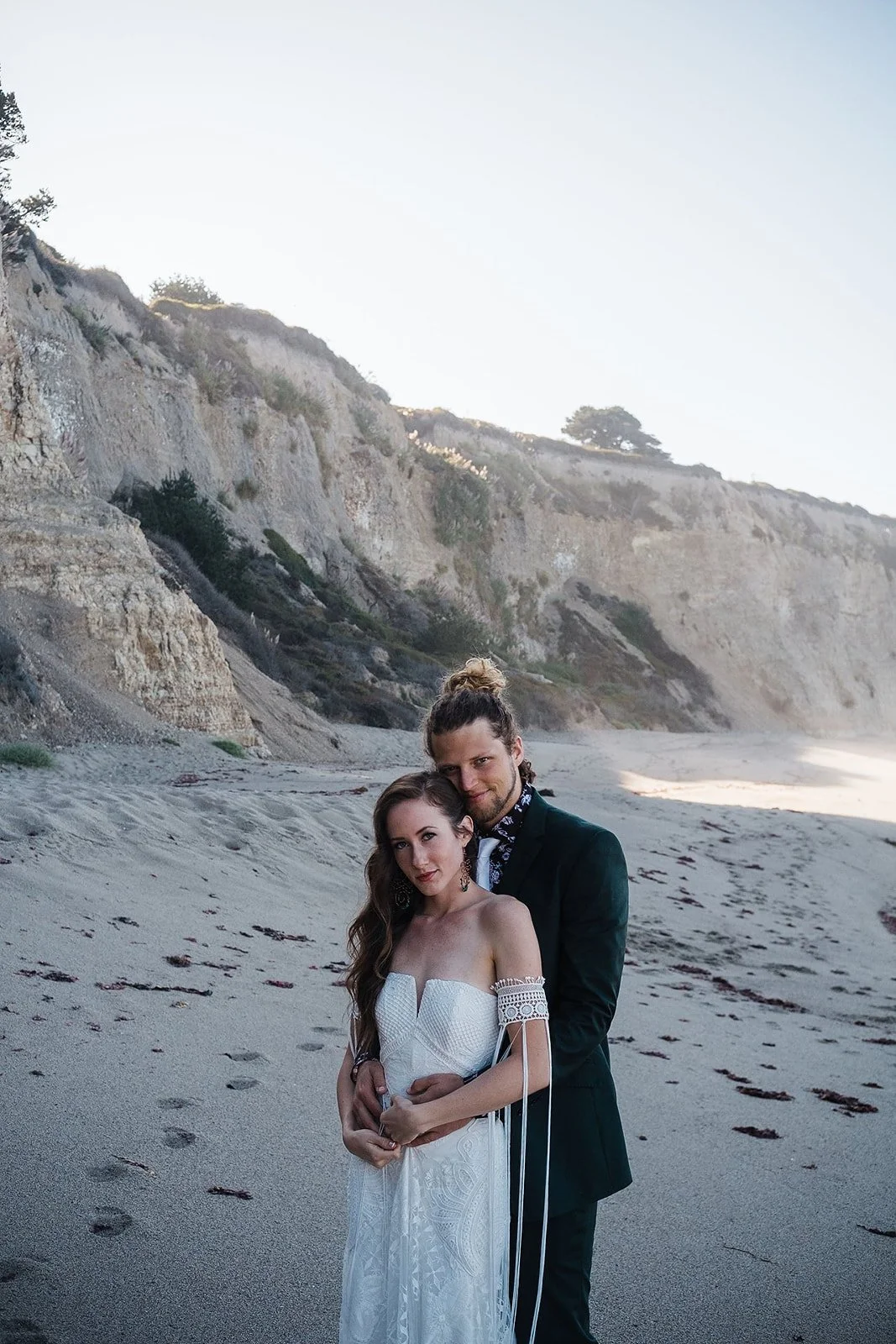 A couple in wedding attire standing on a beach with cliffs in the background. The woman is wearing a white strapless wedding dress, and the man is in a dark suit with a patterned shirt. They are embracing each other, and the woman has long wavy hair,