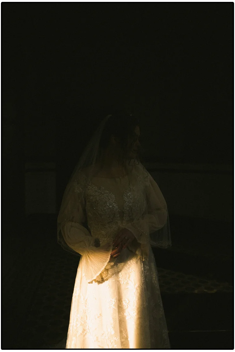 A bride in a wedding dress and veil standing in a dark room illuminated by a single light source, holding a white fan.