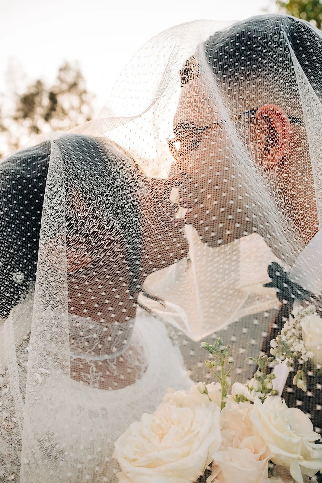 A person wearing glasses and a face mask is partially covered with a lace veil, with flowers in the foreground and blurred trees in the background.