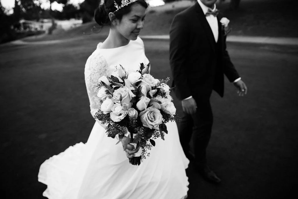 A bride holding a bouquet of roses and greenery, walking with a groom dressed in a tuxedo, outdoors in a park or garden setting.
