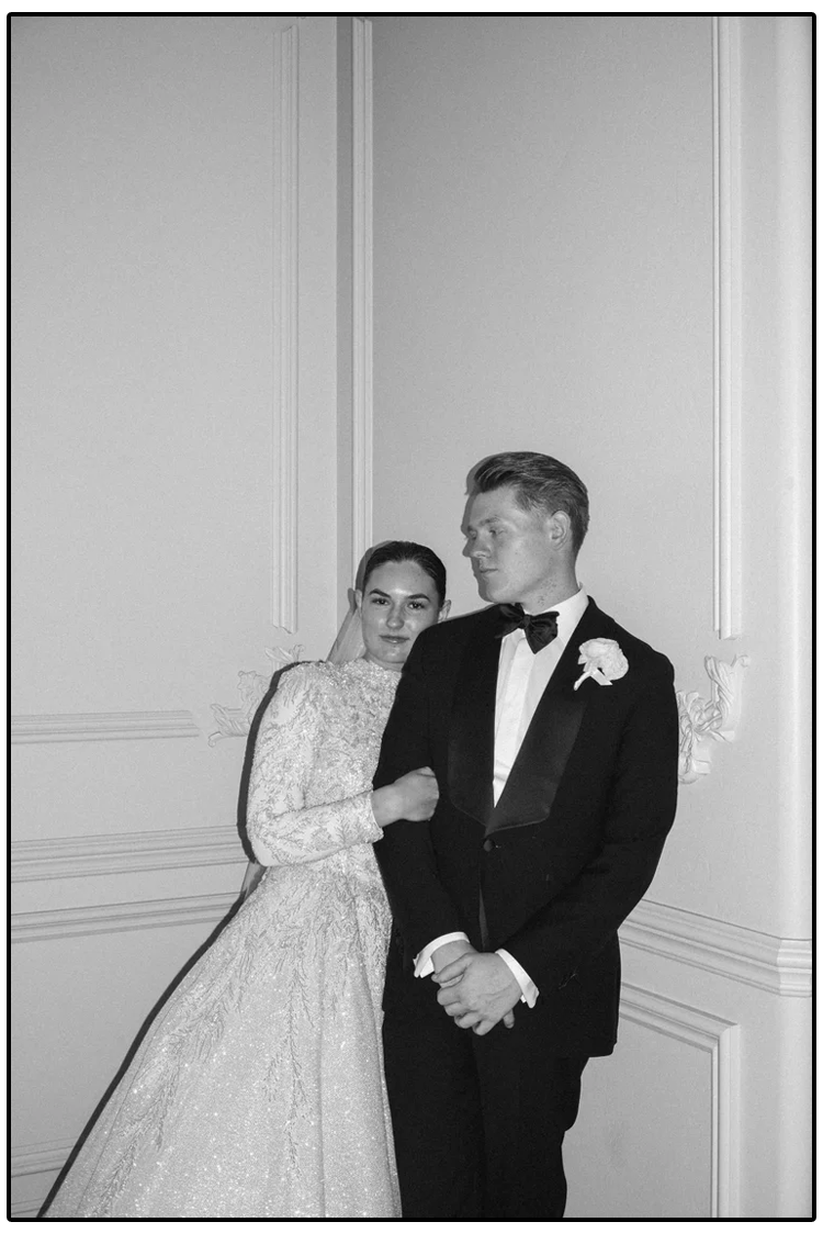 Black and white photo of a bride and groom at their wedding, standing close together; the bride wears a long lace wedding dress and veil, and the groom wears a tuxedo with a bow tie and a boutonniere.