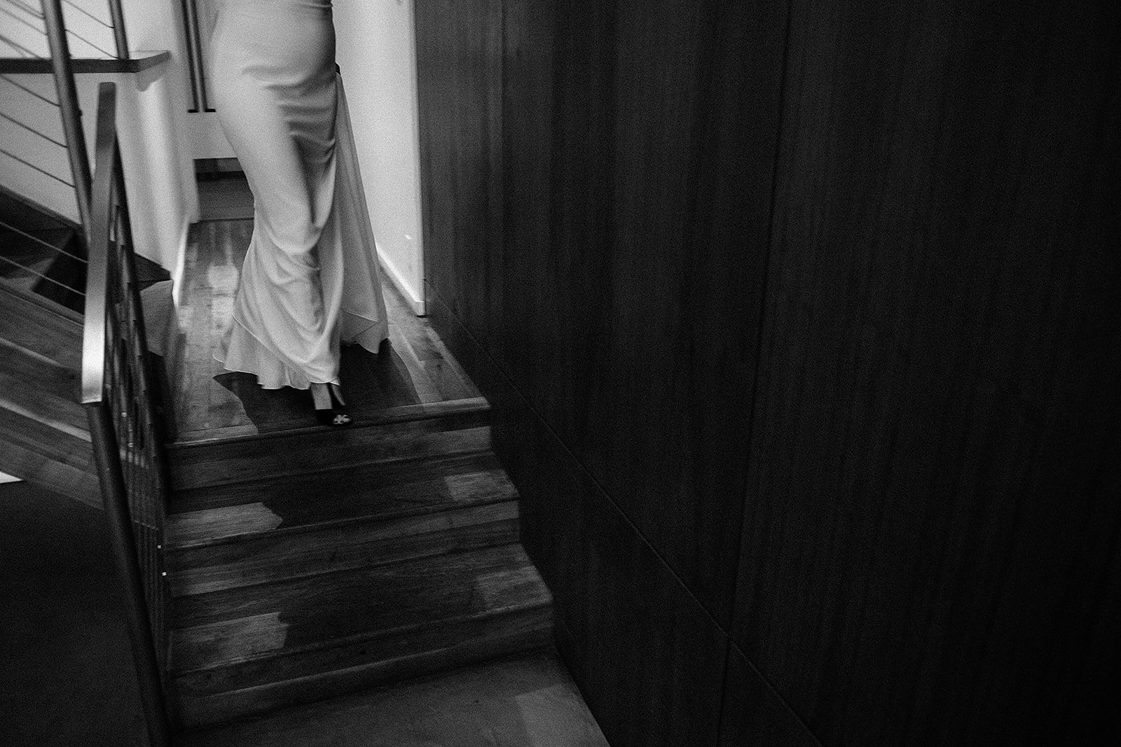 Partial view of a person standing on a small staircase, wearing high heels and a long dress, seen from the back, next to dark wooden wall panels.