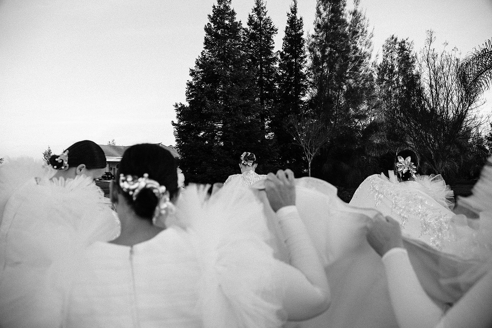 Black and white photo of women dressed in traditional Korean hanbok, sitting outdoors with back facing the camera. They are facing a woman in the focal point wearing a hanbok. Tall trees and a house in the background.