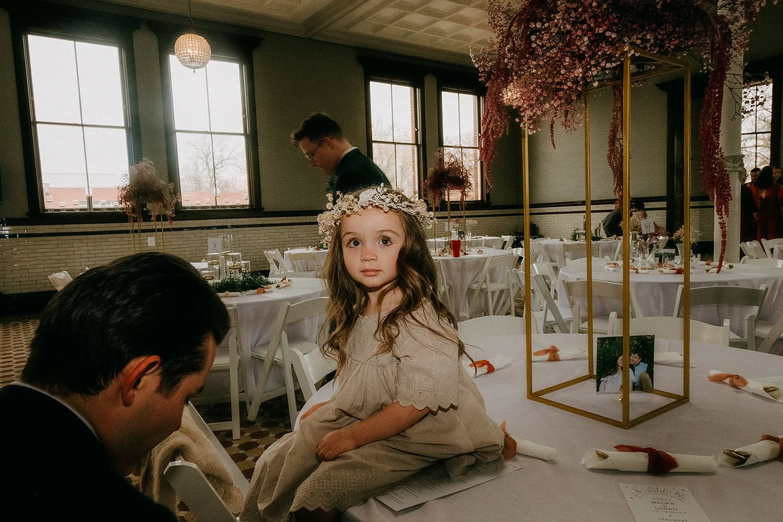 A young girl with long wavy hair, wearing a cream dress and a floral crown, sitting on a table in a decorated event space with white tablecloths and floral centerpieces. People are in the background inside a room with large windows and hanging pink f