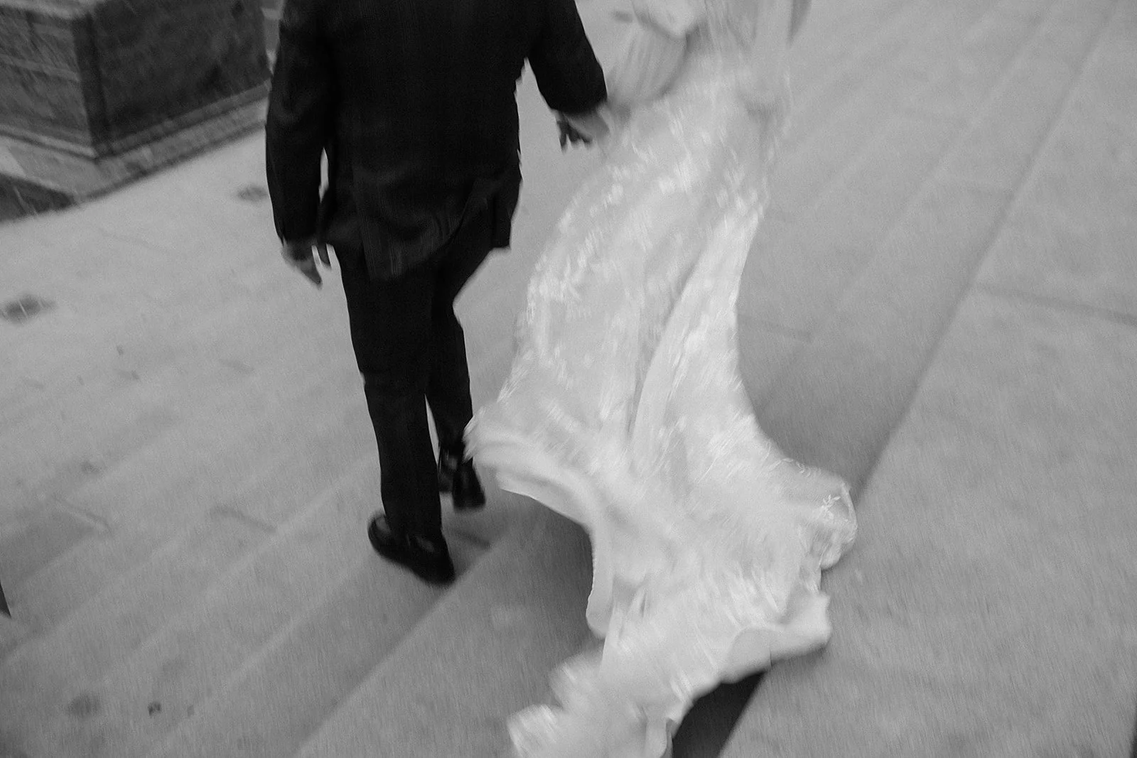 A person dressed in a dark suit, walking on a sidewalk, holding a large piece of fabric or material that is draped on the ground.