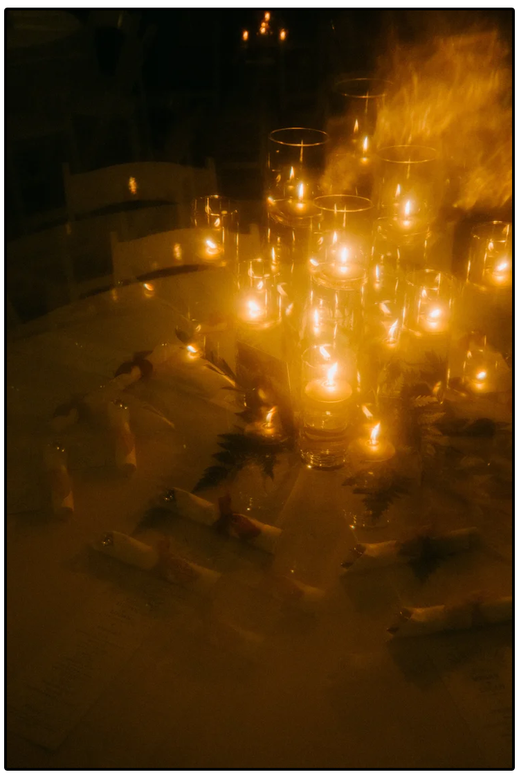 A table decorated with several lit candles and small wrapped gifts, with chairs around it, in a dimly lit setting.