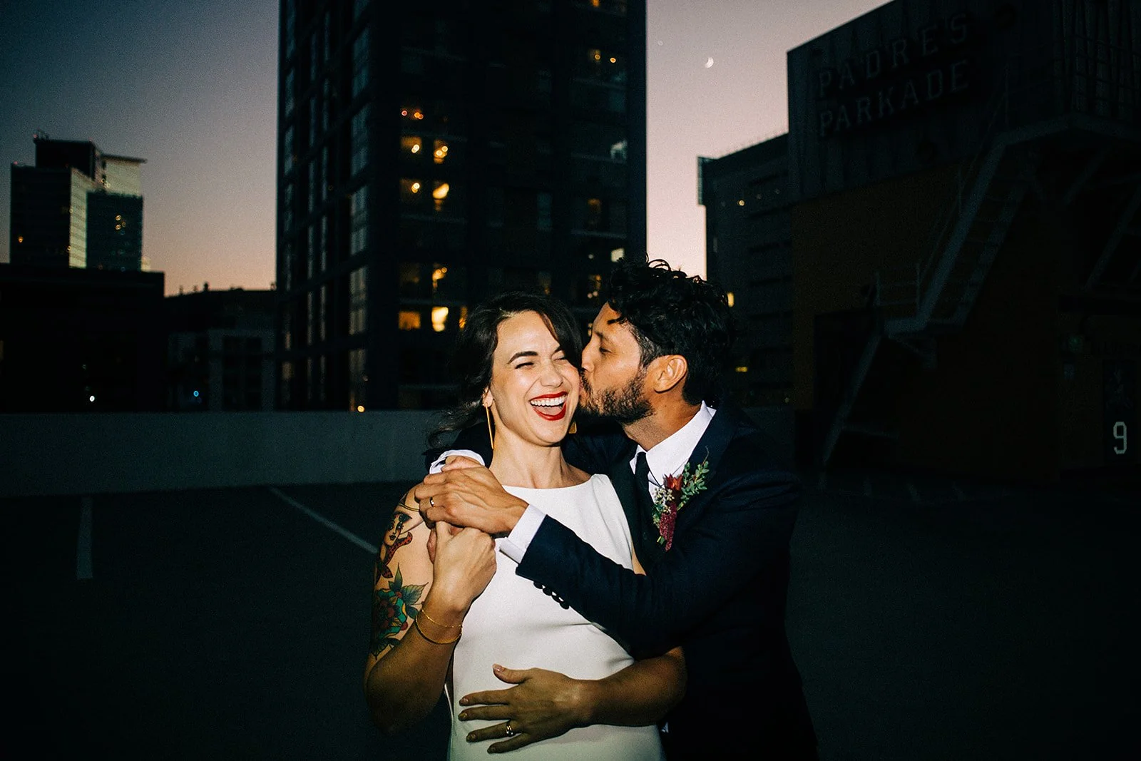 A joyful couple sharing a dance on a rooftop at dusk in an urban setting, with the city skyline and buildings in the background.