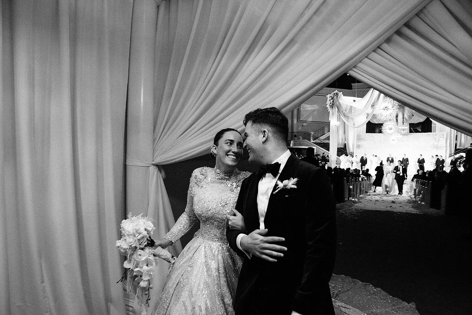 A bride and groom smiling together at their wedding reception, seen through a curtain with wedding guests and a decorated stage in the background.