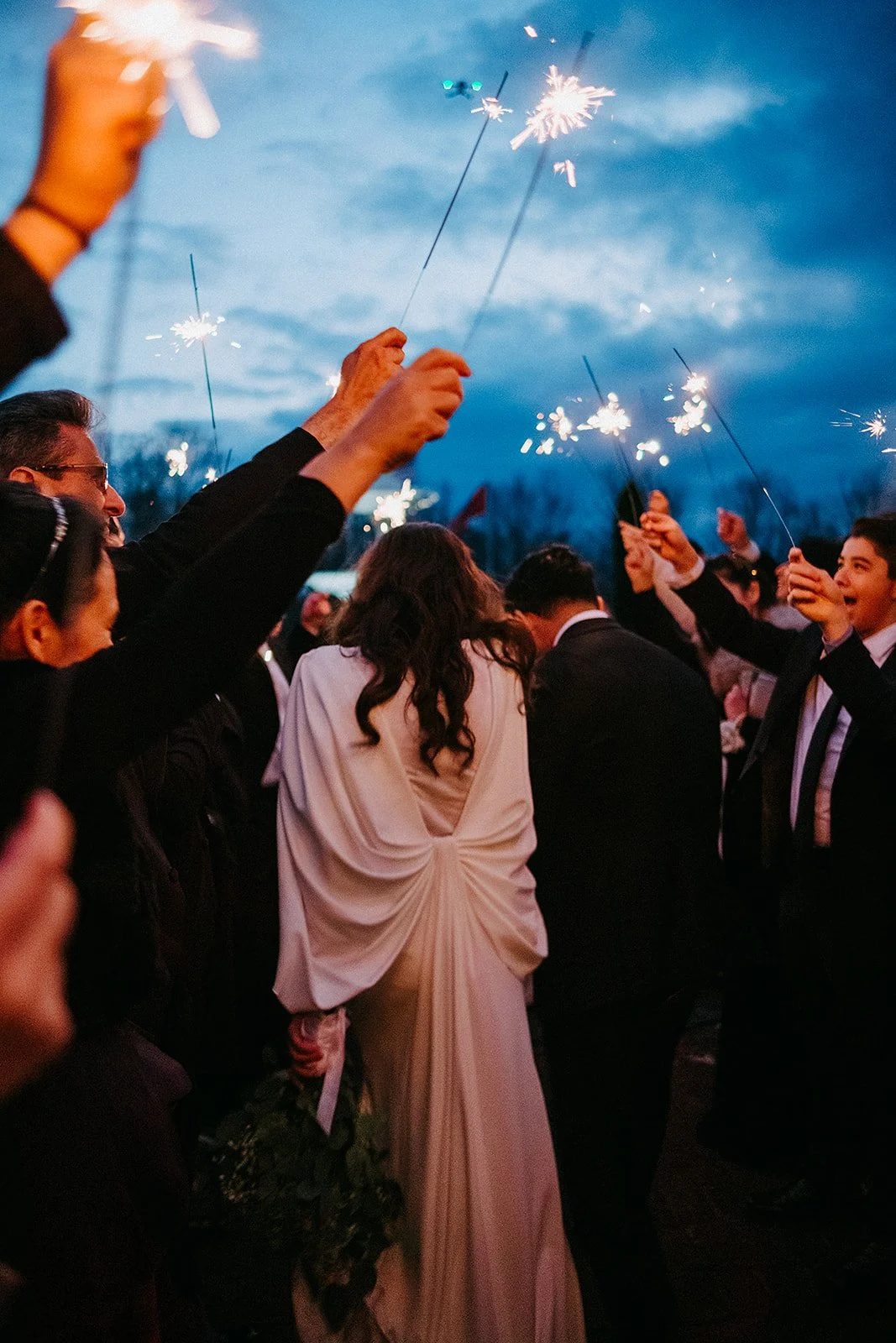 People celebrating outdoors at sunset, holding sparklers in the air, with the sky turning blue.