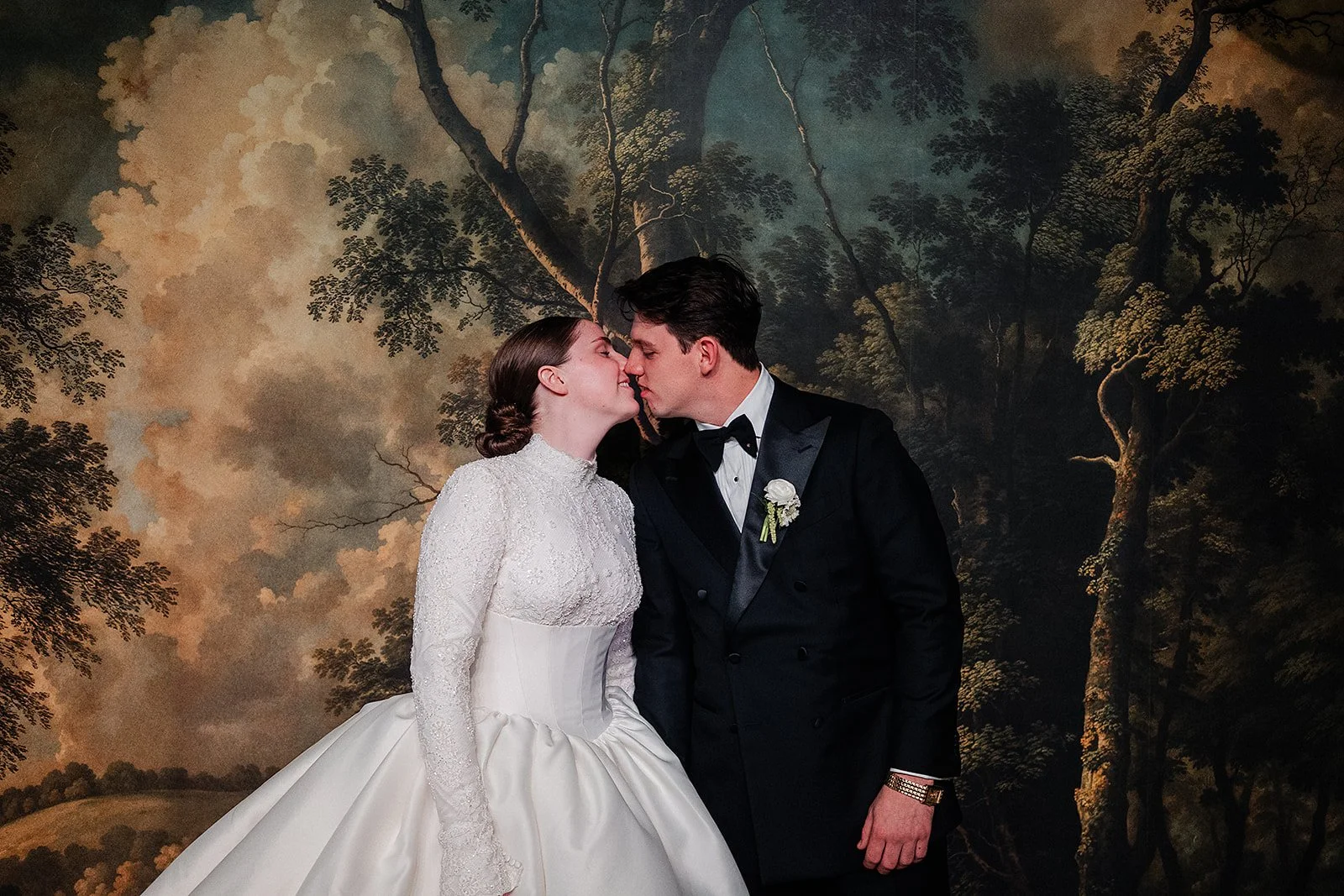 A bride and groom are about to kiss, standing closely with their foreheads touching. The bride is wearing a white wedding dress with lace details, and the groom is dressed in a black tuxedo with a white shirt and black bow tie. The background feature