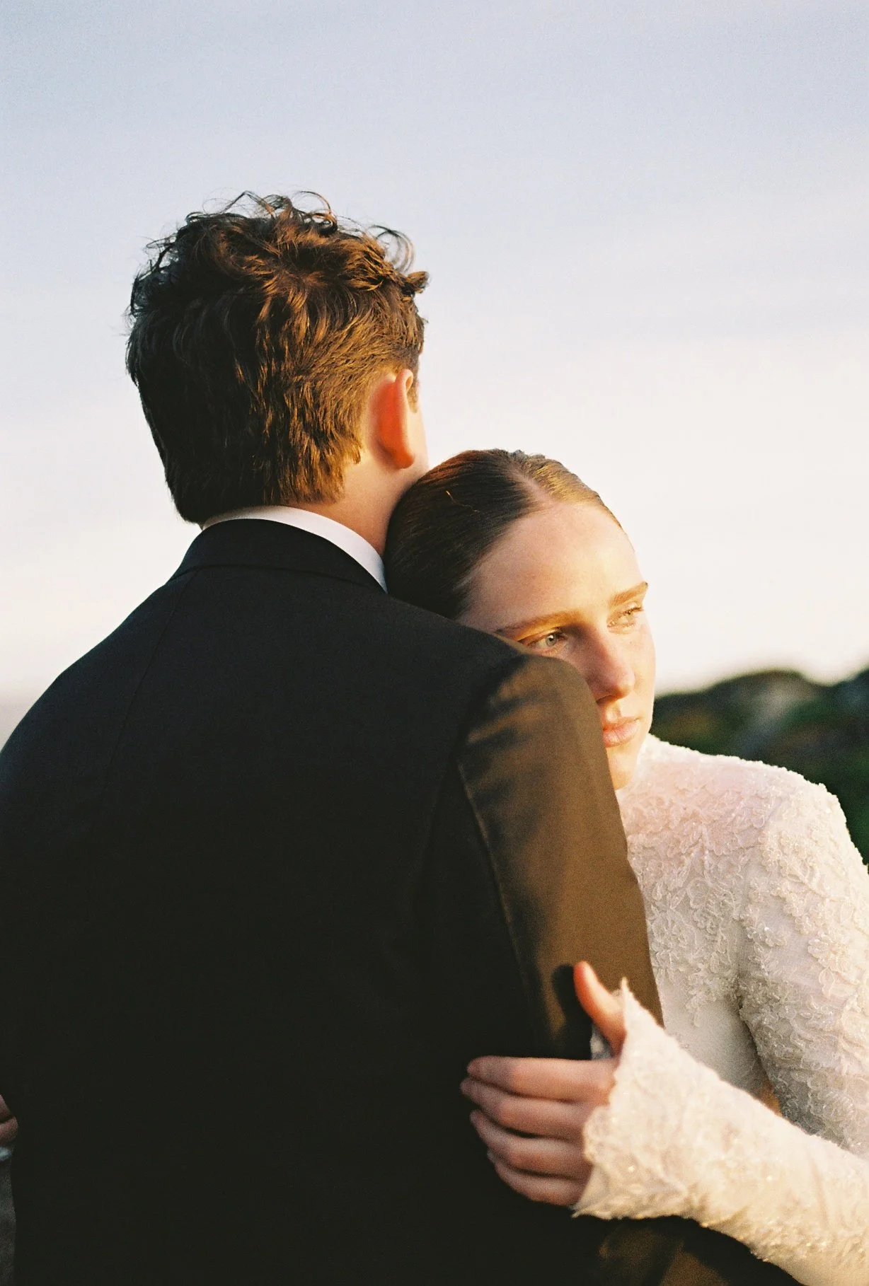 A man with curly hair and a dark suit embraces a woman with brown hair in a white lace wedding dress, both looking toward the right, against a softly lit outdoor background.