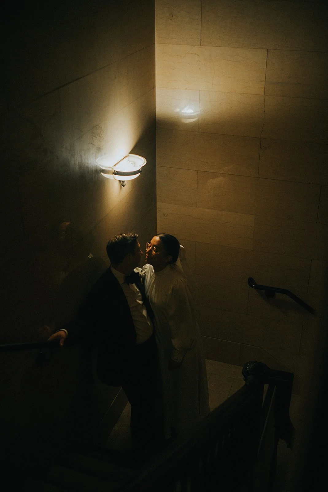 A couple dressed in formal attire standing on a staircase in a dimly lit indoor setting, with a wall-mounted lamp casting warm light.