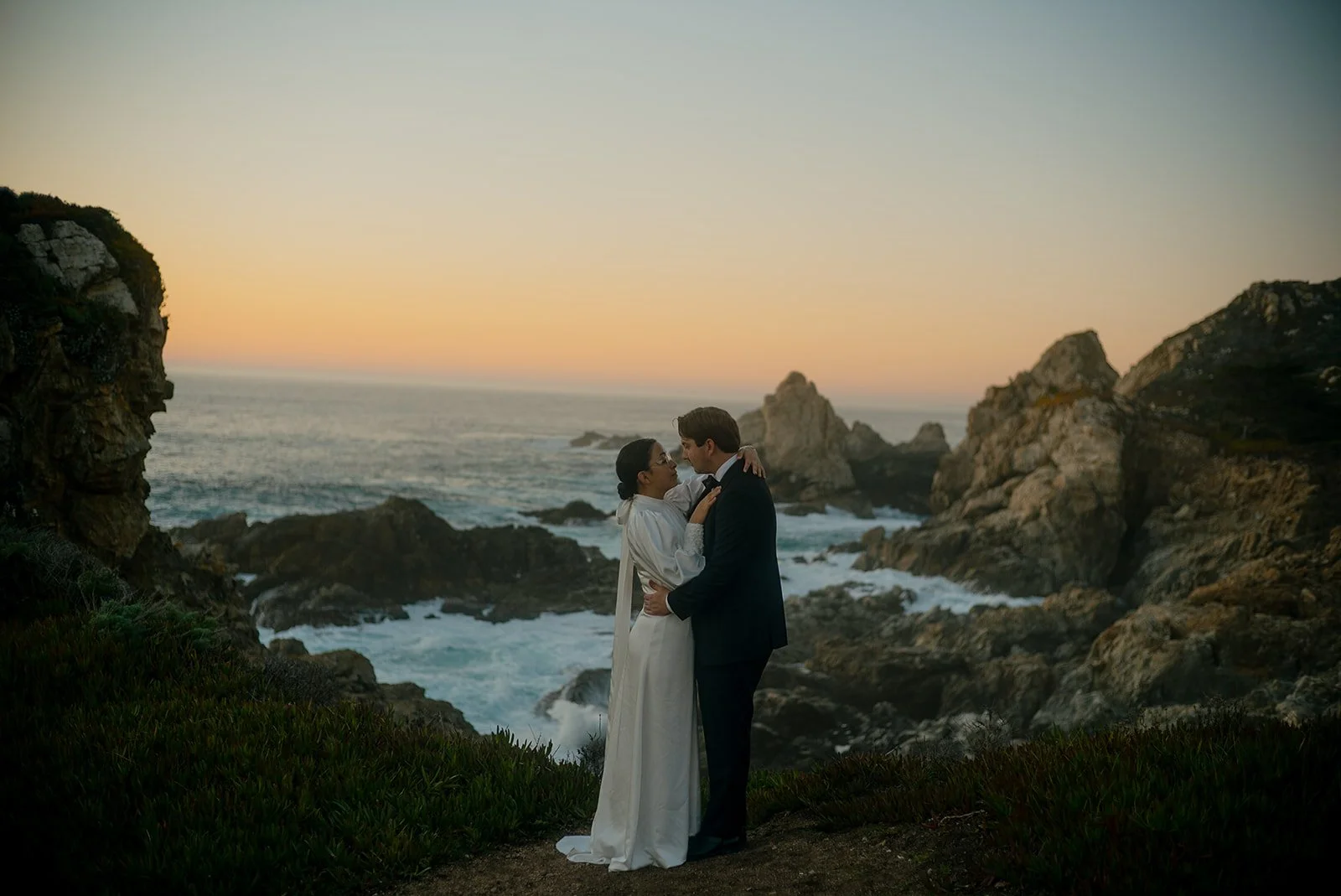 A couple dressed in formal wedding attire embracing on a rocky coastline at sunset, with the ocean and cliffs in the background.