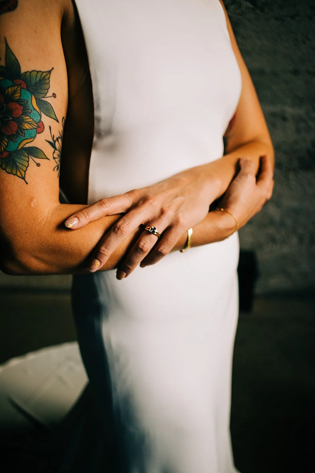 Close-up of a woman with a colorful floral tattoo on her left arm, wearing a white dress and jewelry, hands crossed in front of her, standing on a dark floor.