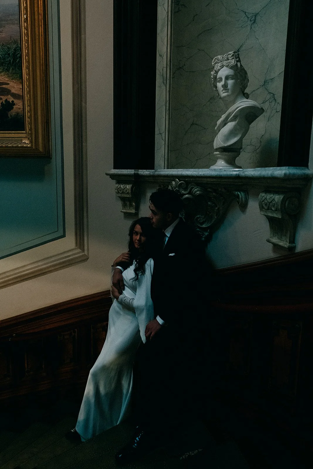A man and woman in formal attire standing next to each other in a dimly lit gallery or museum, with a portrait bust sculpture of a woman in the background and framed painting on the wall.