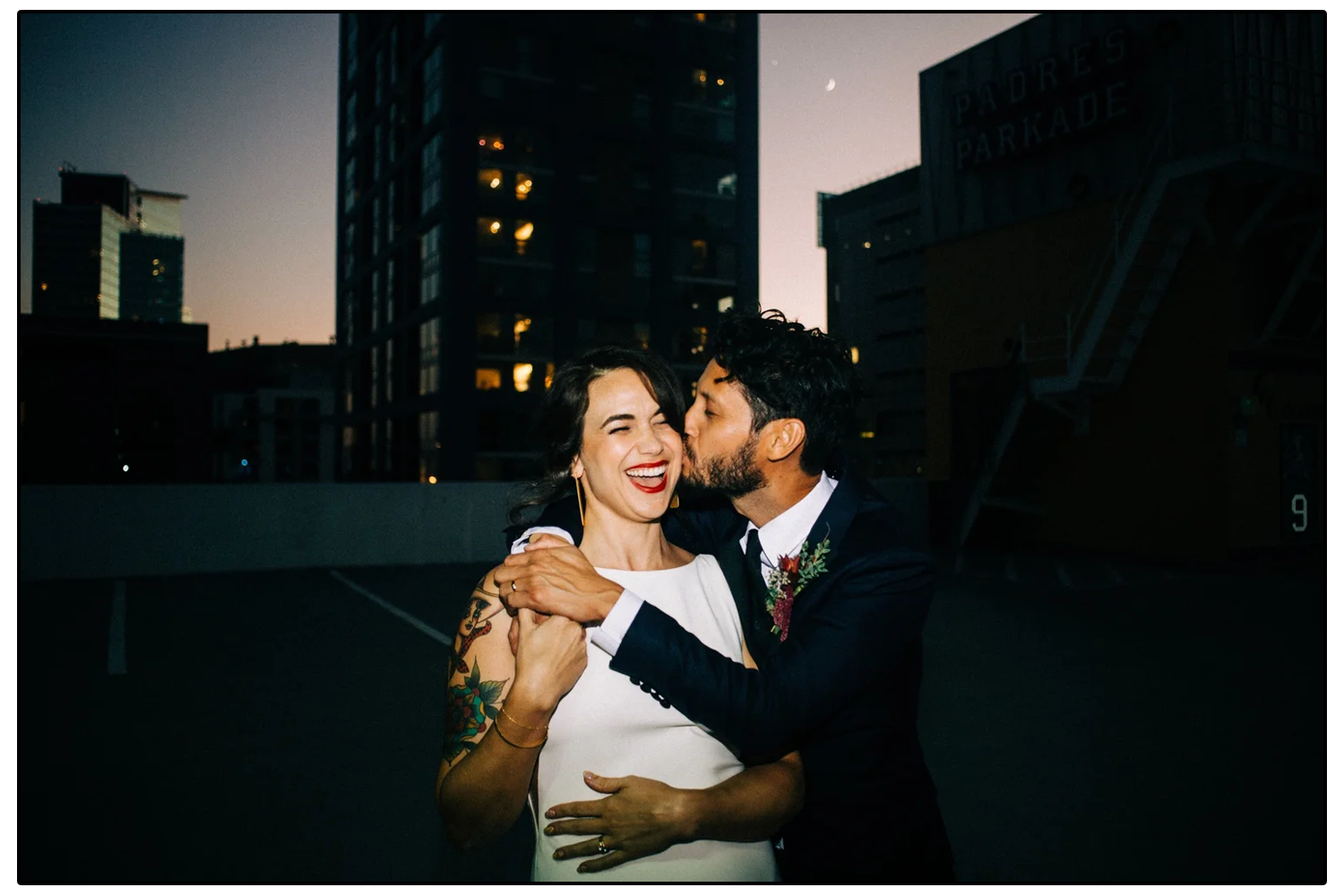 A happy couple in wedding attire sharing a kiss on a city rooftop at dusk, with tall buildings in the background.
