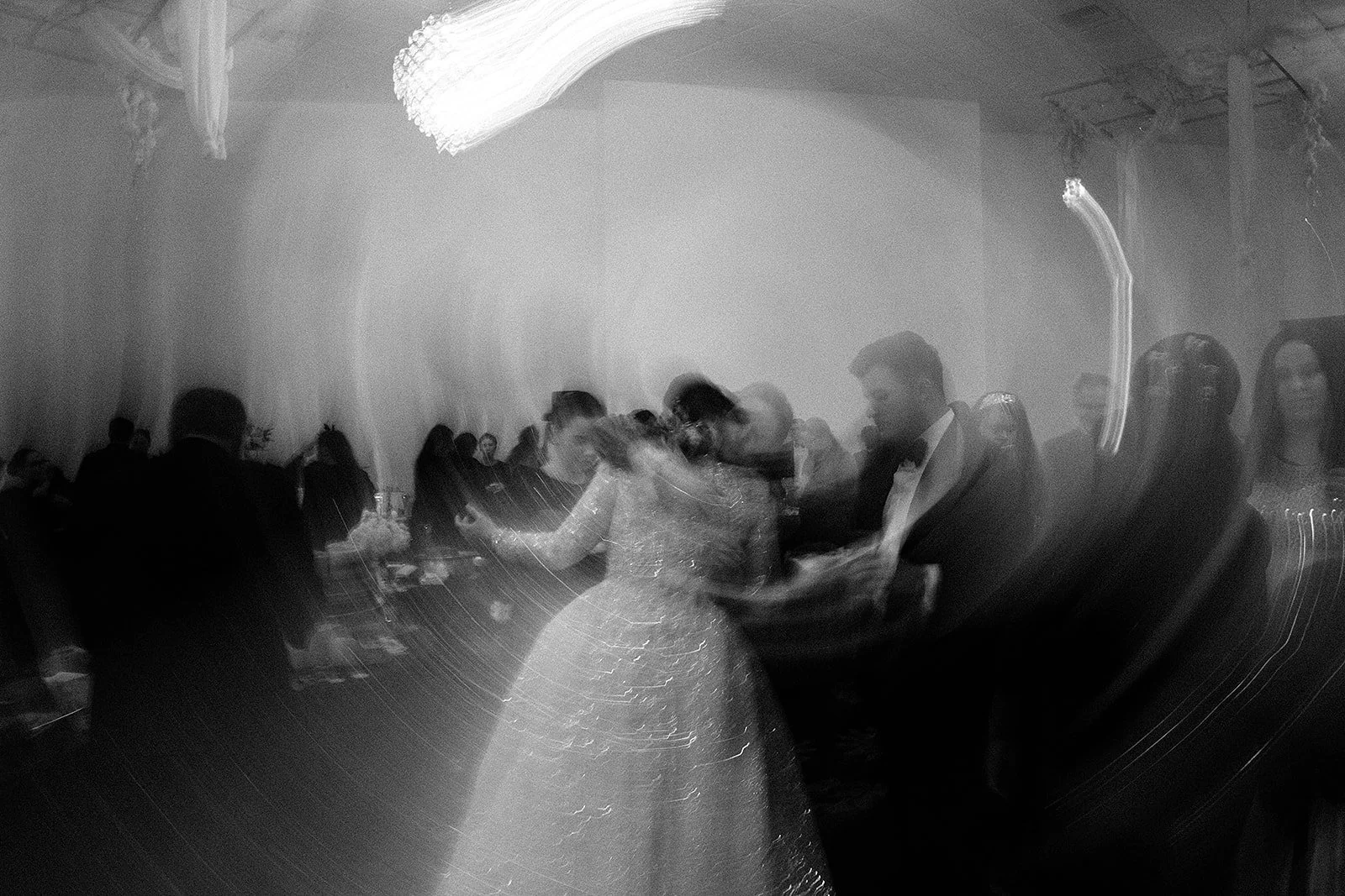 People dancing and socializing at a formal event with a large chandelier, in black and white with light streaks.
