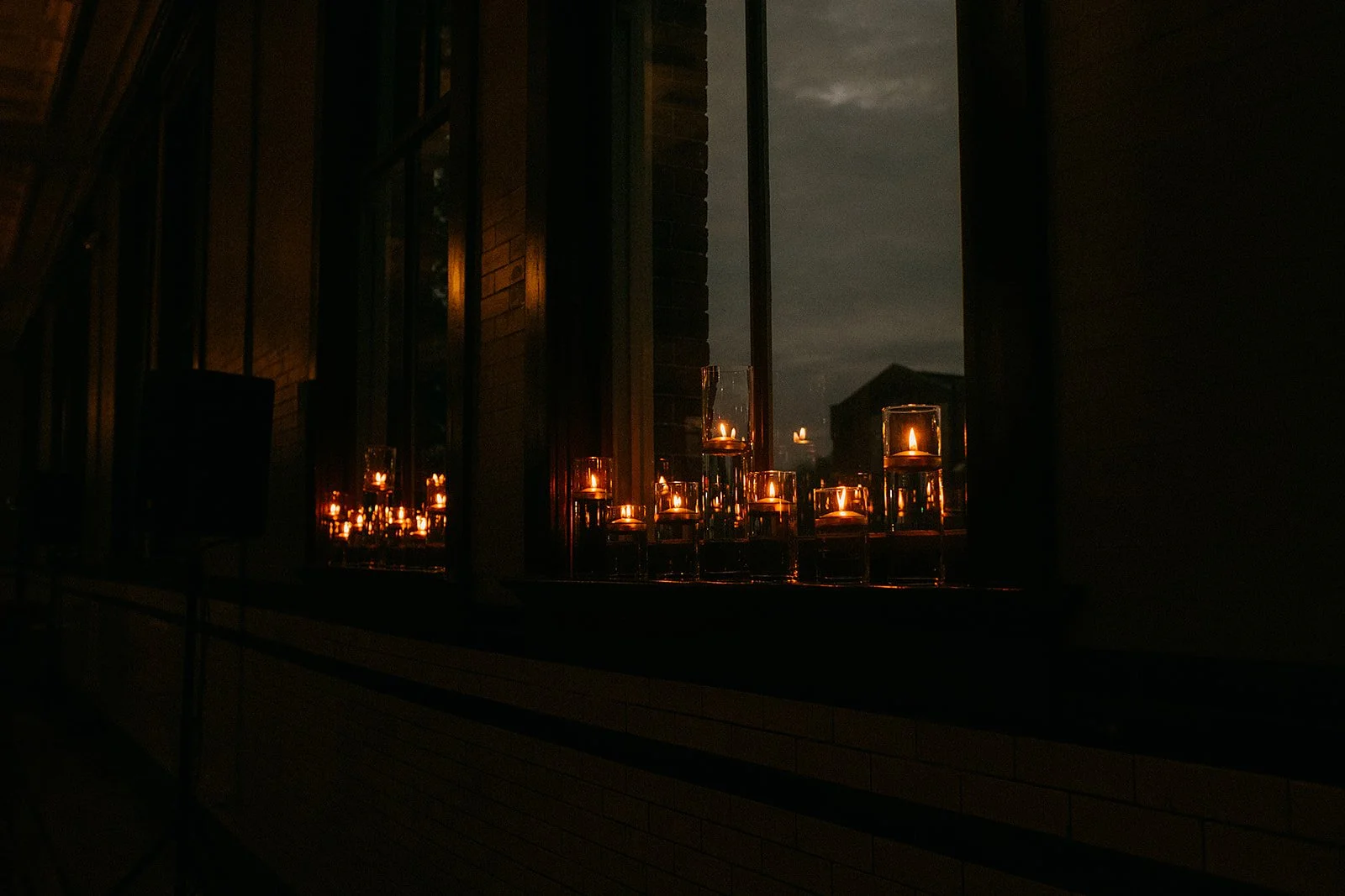 Nighttime view of candles in glass holders on a window sill, with a dark sky and silhouette of a building outside.