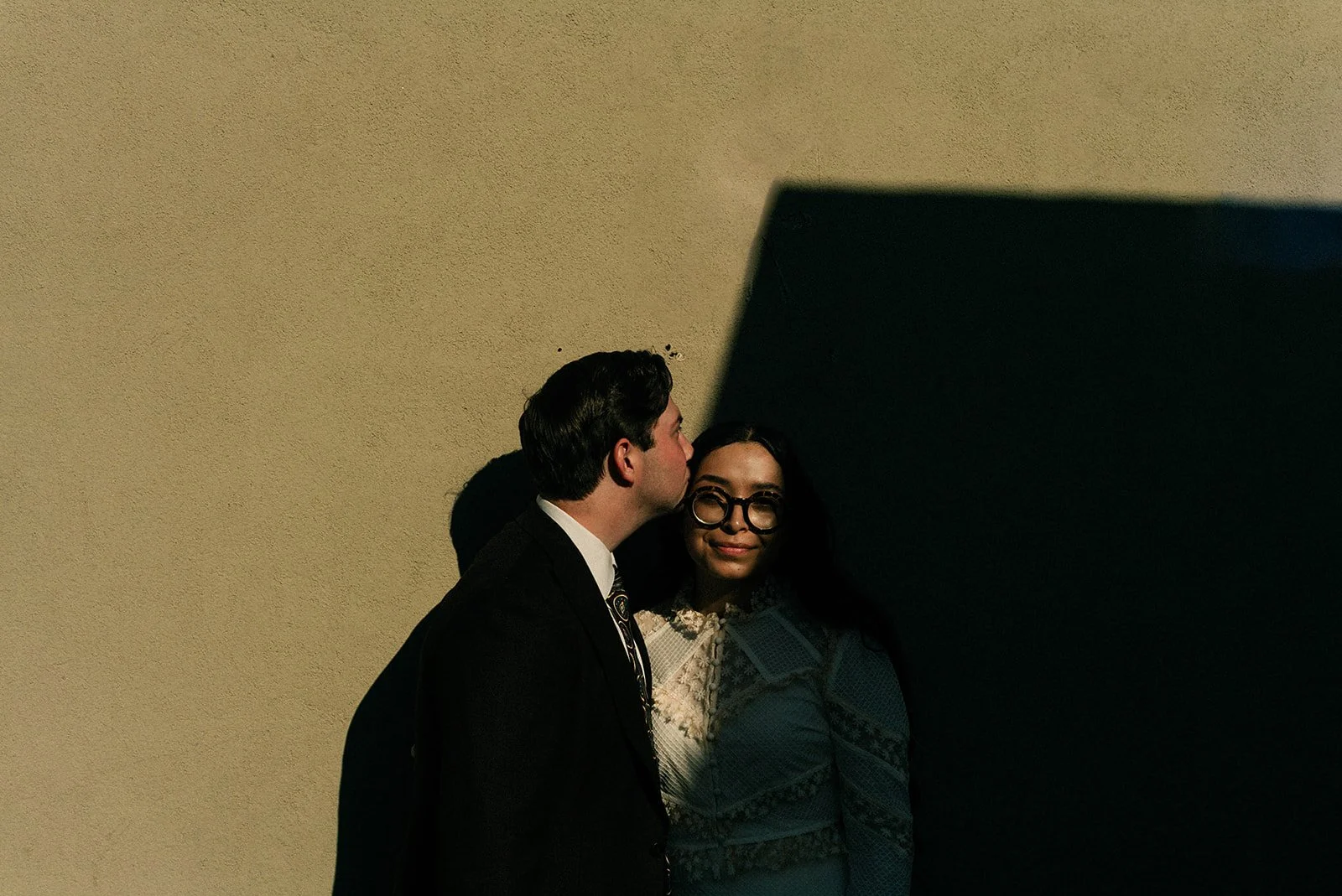 A man in a suit kisses a woman in glasses on the cheek against a beige wall, with a dark shadow partially covering her.