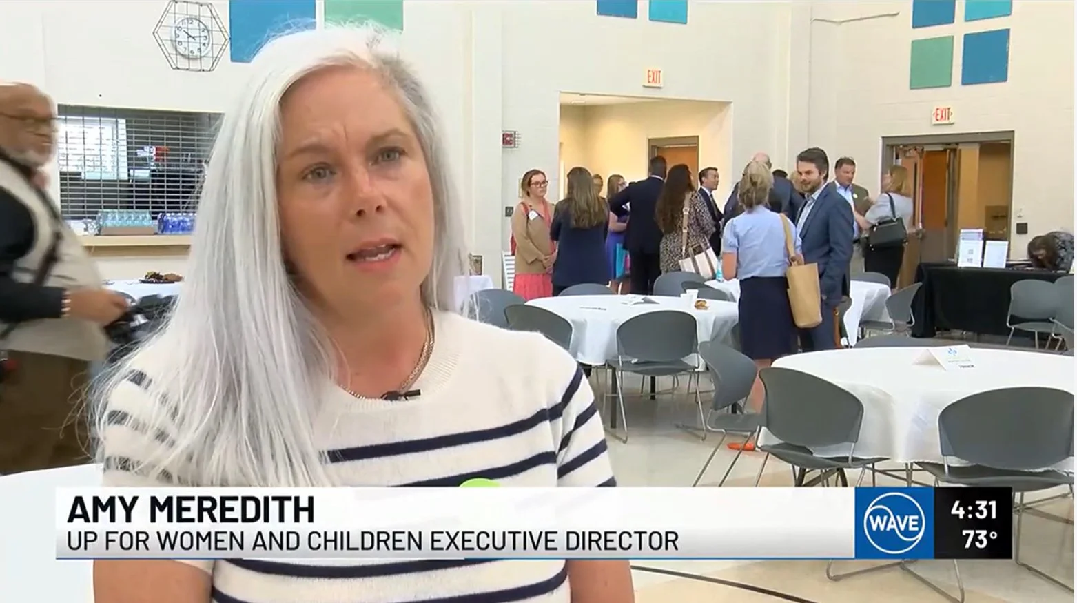 Amy Meredith, UP for Women and Children Executive Director, speaking at an indoor event with white round tables and chairs, and groups of people standing and conversing in the background.