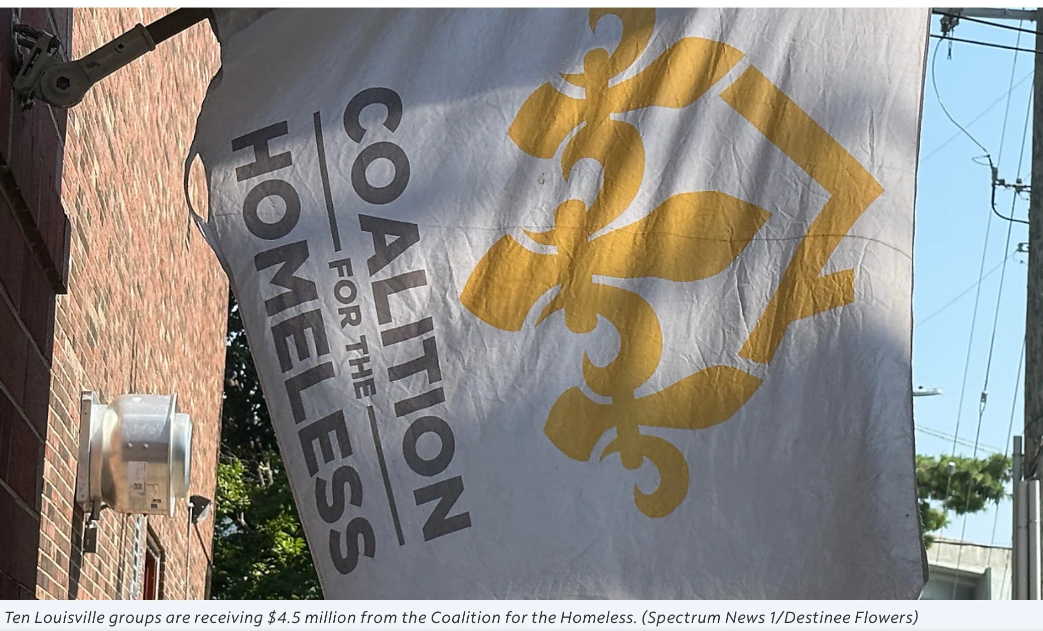 Flag with gold fleur-de-lis and text reading "Alliance for the Homeless" hanging outside a brick building, with power lines and some greenery in the background.