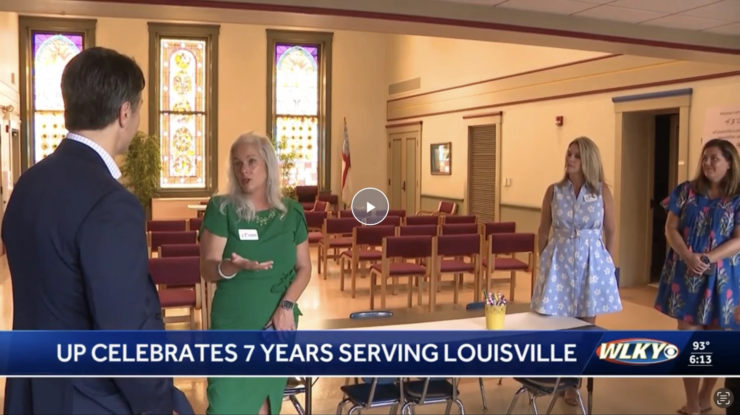 Women having a conversation with a man inside a church or community hall with stained glass windows, chairs, and an American flag in the background.